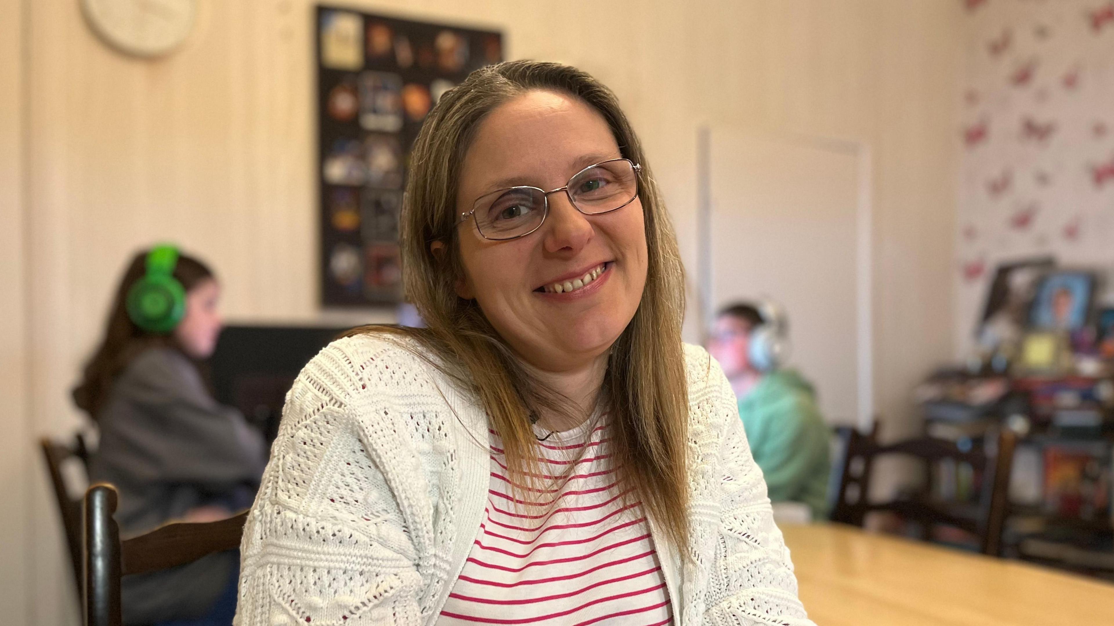 Liam's mum Elena smiles directly at the camera. She has mid-length blonde hair and is wearing silver framed glasses. She is wearing a red and white striped top with a white cardigan. In the background (although blurred) - Liam and Aidan can be seen playing on their computers. 