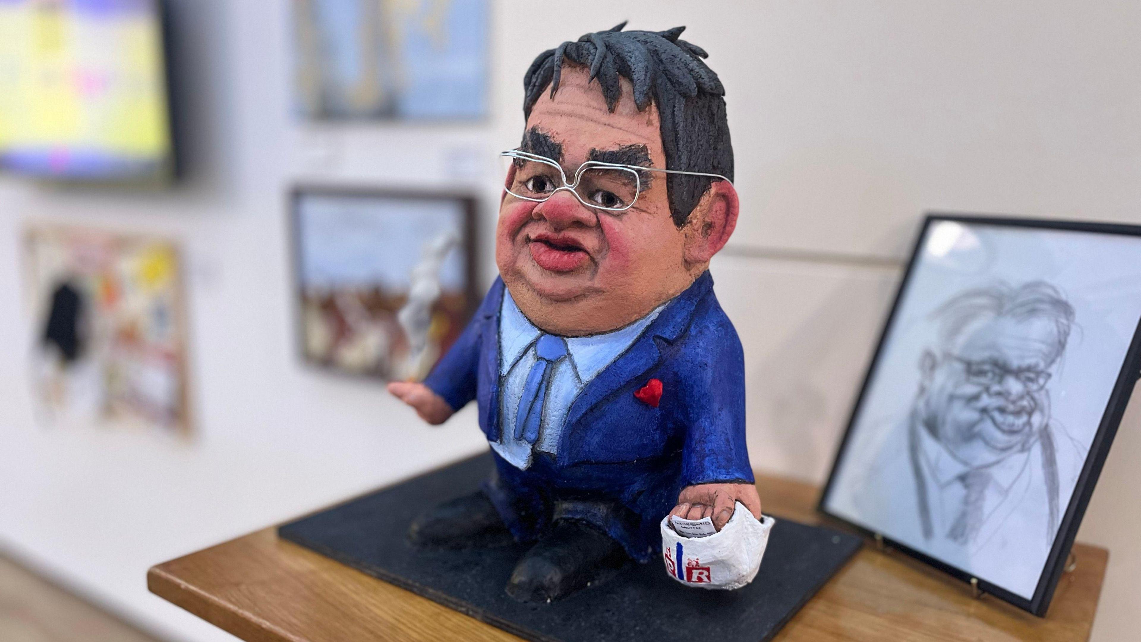 A papier mâché caricature of Guernsey deputy John Gollop, with similarly grotesque pencil sketch behind. He is wearing an ill-fitting blue suit with his shirt untucked. He wears glasses and holds a plastic carrier bag stuffed with papers in one hand and a lit cigarette in the other.