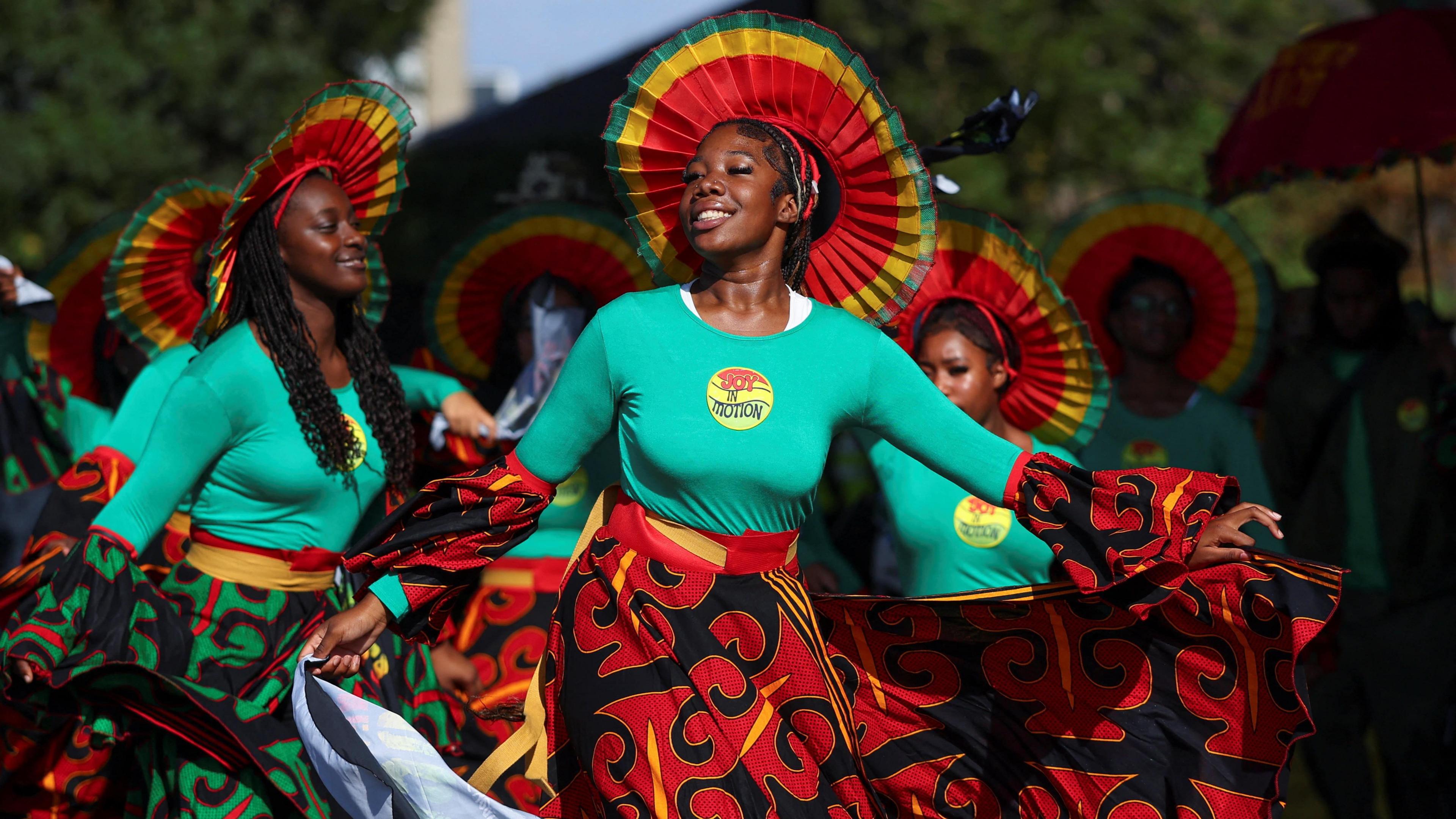 A group of women in green tops and patterned skirts of red and green dance through the street. They wear fan-like headpieces with red, yellow and green stripes.