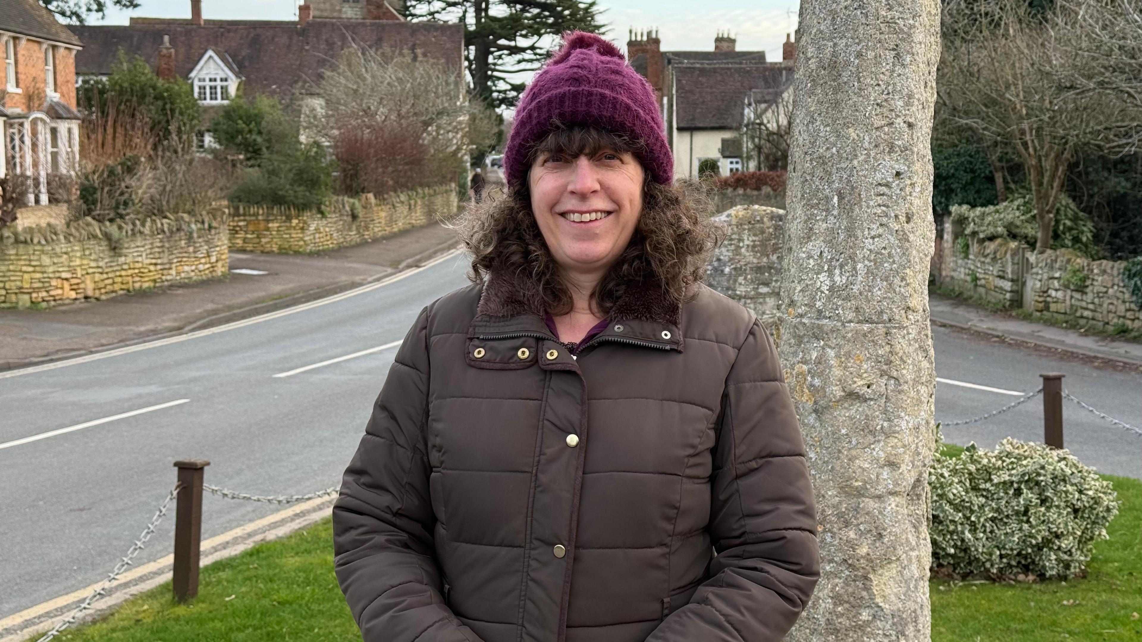 A woman with a maroon hat and brown jacket stands and smiles at the camera.