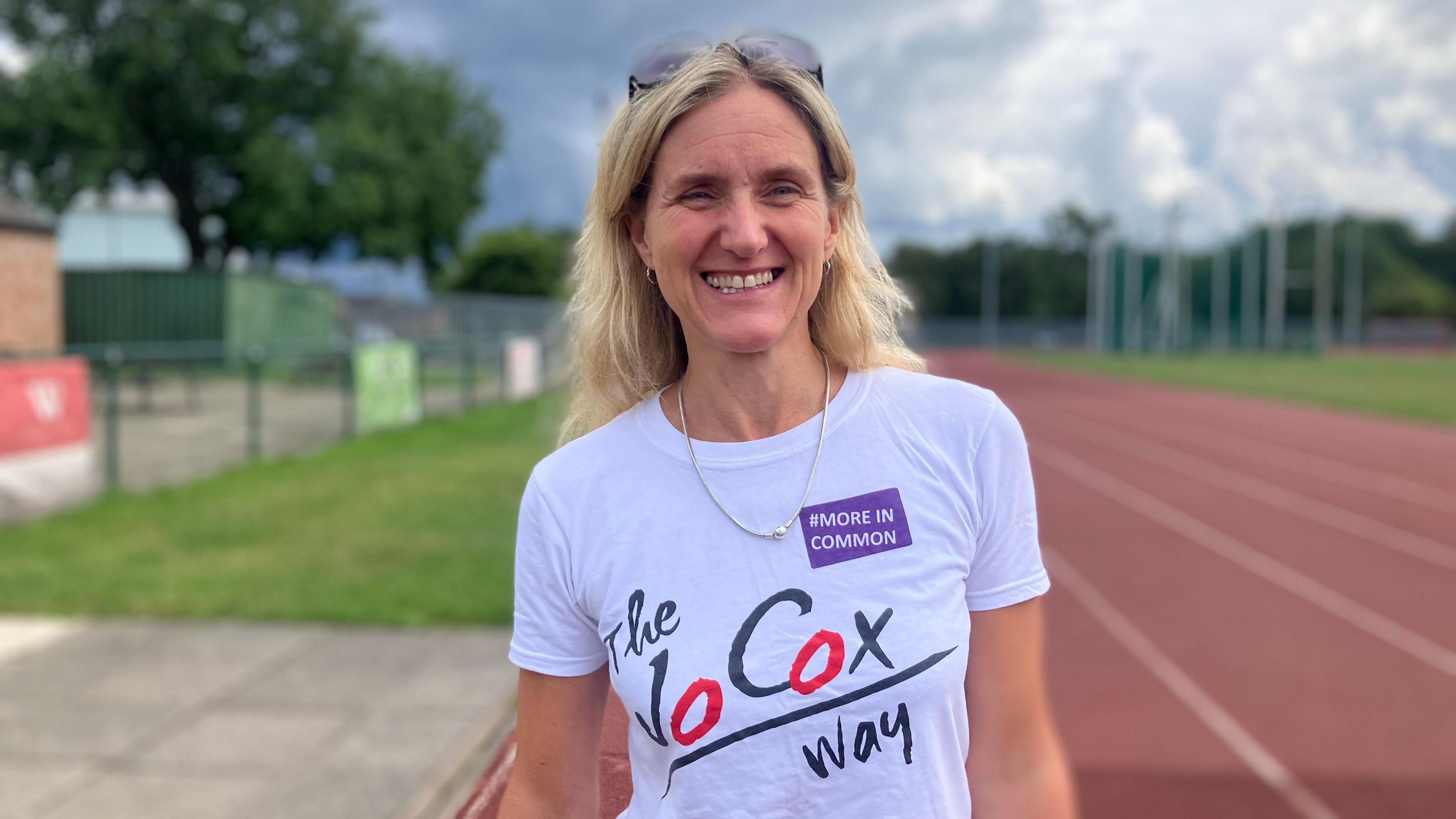 Kim Leadbeater MP, a blonde woman, smiles at the camera. She wears a white top with "The Jo Cox Way" on the front. She wears a silver chain with sunglasses on the top of her head. Behind her a race track is blurred.
