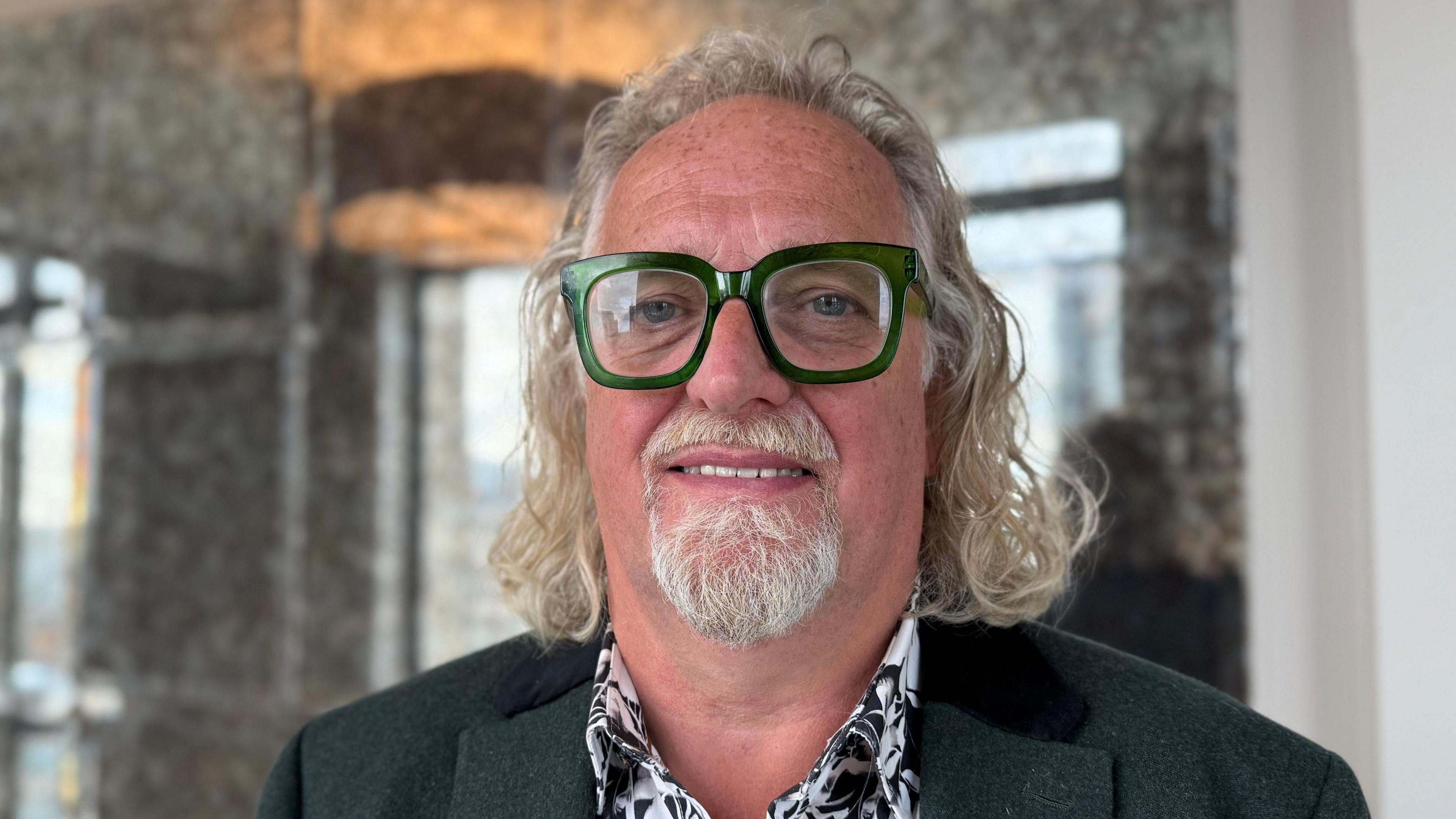 Michael Stewart, a man with long grey hair, a grey beard, green glasses and a green jacket. He has a flowery shirt.