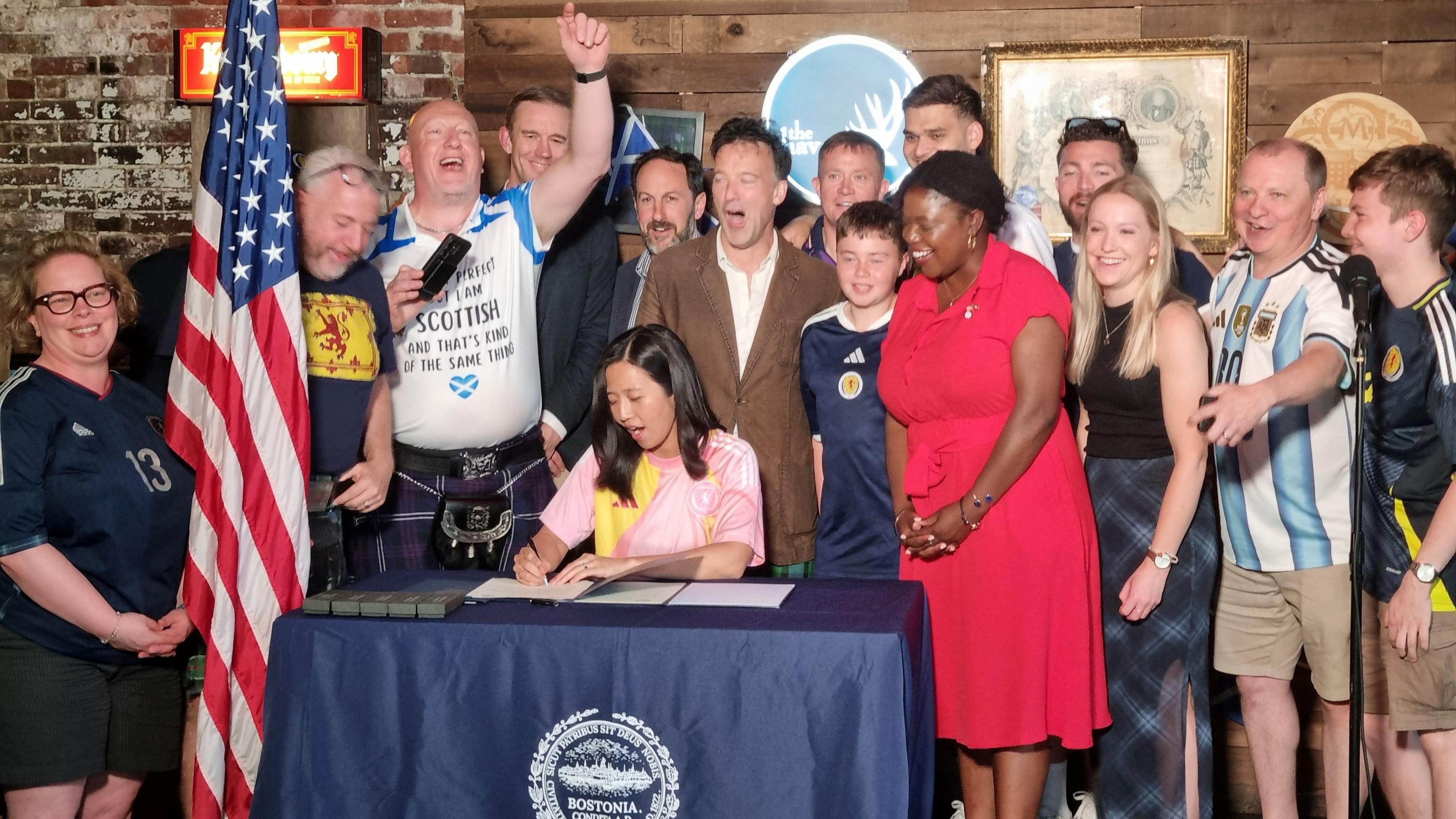 Mayor of Boston Michelle Wu signing a document in a Scottish pub in Boston. She is surrounded by people