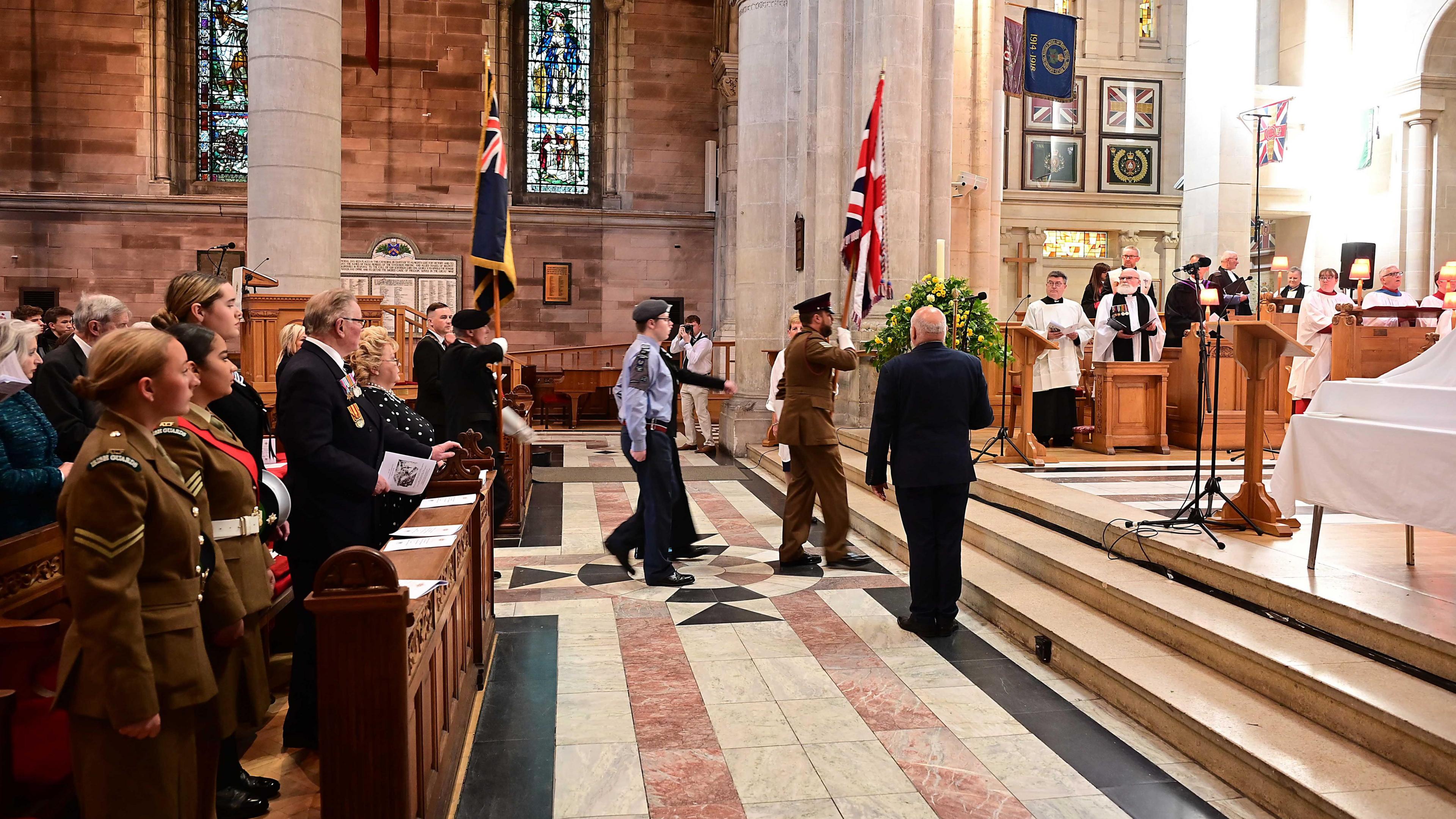 A wide shot of the interior of Belfast Cathedral. In the rows, those in military uniforms watch on as a Union Jack is brought up to the alter of the Cathedral by a uniformed officer. To the side are clergy representatives in white and black robes. The cathedral is made up of white stone walls and marbled forced featuring white, pink and black squares.
