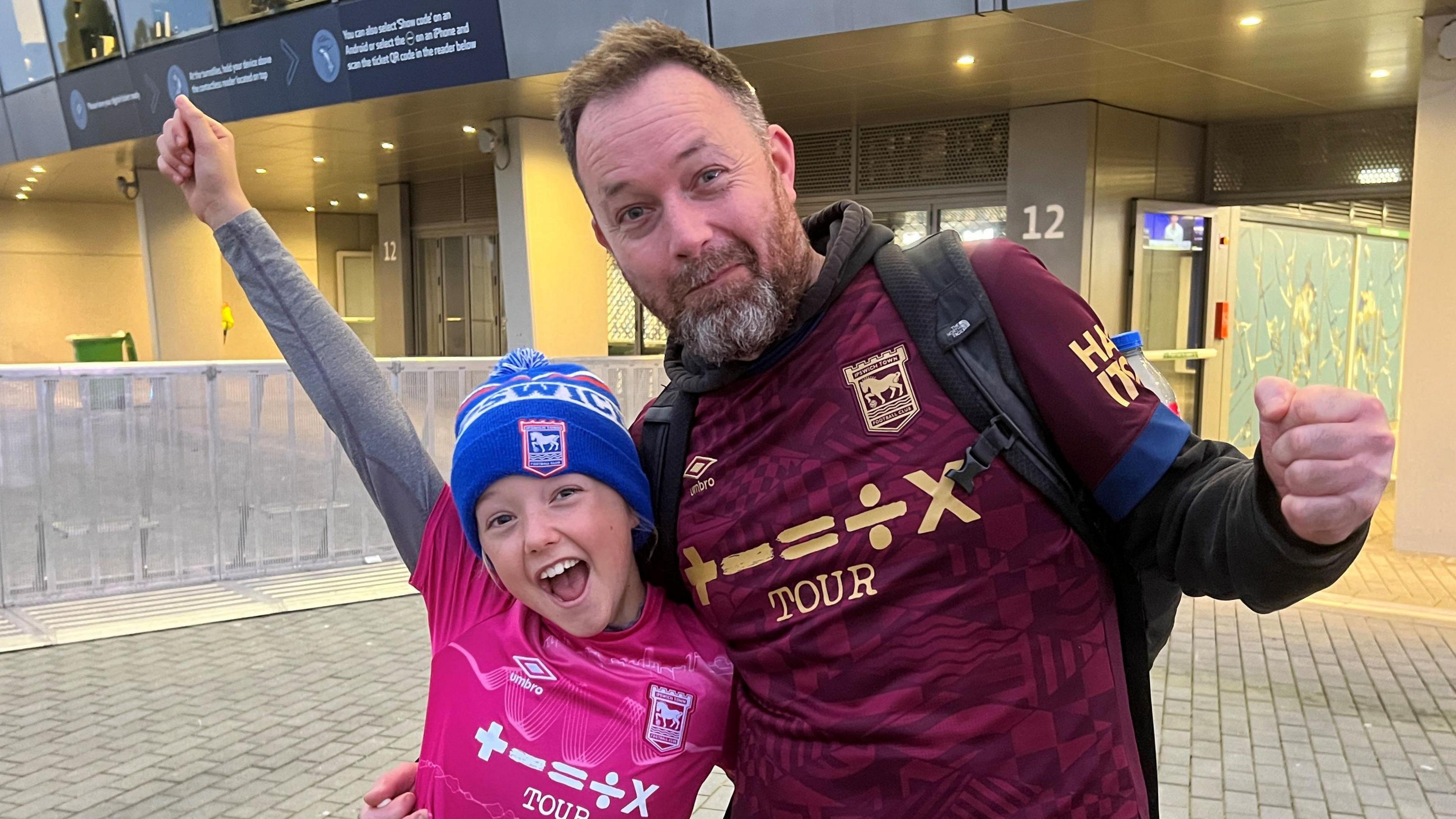 A father and daughter are in the centre of the picture.  The dad has short brown hair with a greying, neat beard and moustache.  He's wearing the maroon Ipswich Town 2024 shirt with the Ed Sheeran sponsor slogan - he's leaning into his daughter with his right arm around her while his left hand is clenched in a fist. The daughter is wearing the pink third kit Ipswich Town shirt with the Ed Sheeran sponsorship. She has a grey long sleeve top underneath and a blue and white Ipswich Town bobble hat pulled down to her eyebrows.  She has her left arm around her dad's waist while her right arm is raised in victory.  In the background is the Tottenham Hotspur Stadium.