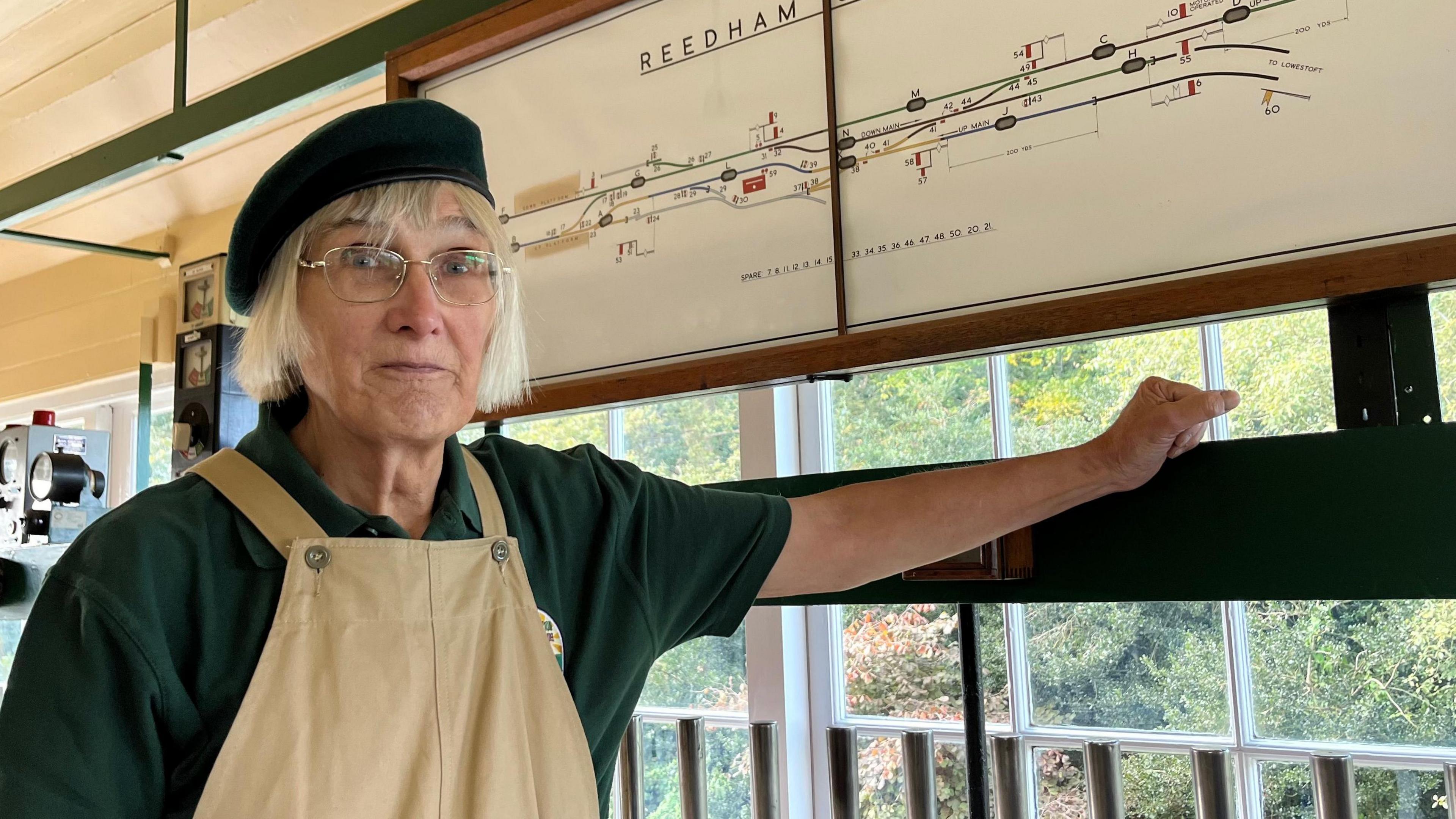 Michelle Davidson with a short blonde bob and has gold rimmed glasses on and a green hat. She is standing in front of a Reedham junction railway line sign and is wearing a green polo shirt and brown overalls. 