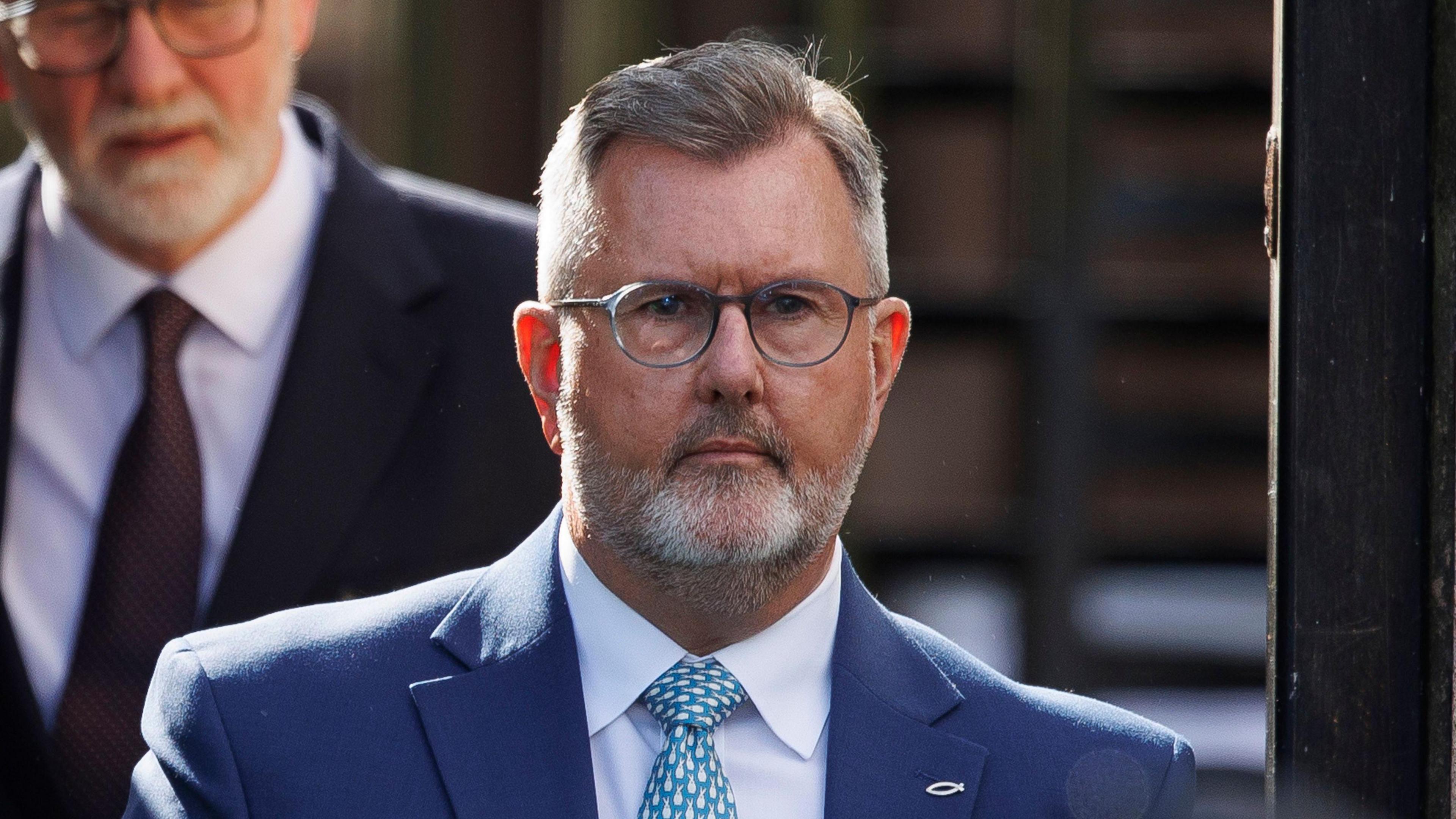 Sir Jeffrey Donaldson, pictured from the shoulders up, wearing a dark blue suit, light blue shirt and spotty blue tie. He has a tight grey beard and grey hair and is wearing glasses. 