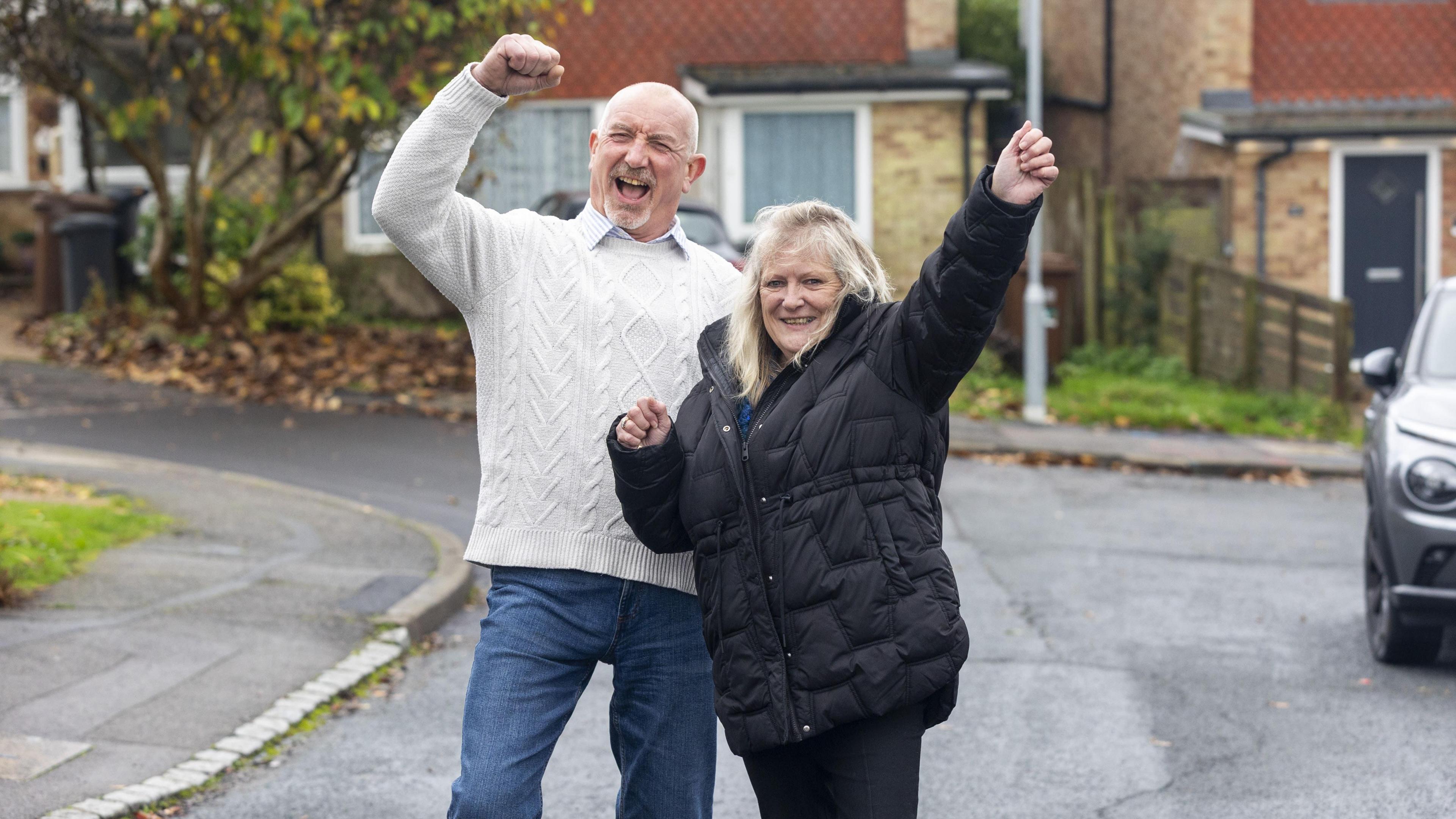 Man in white jumper and woman in dark coat both with hands in the air in celebration stading in a cul de sac with houses behind them