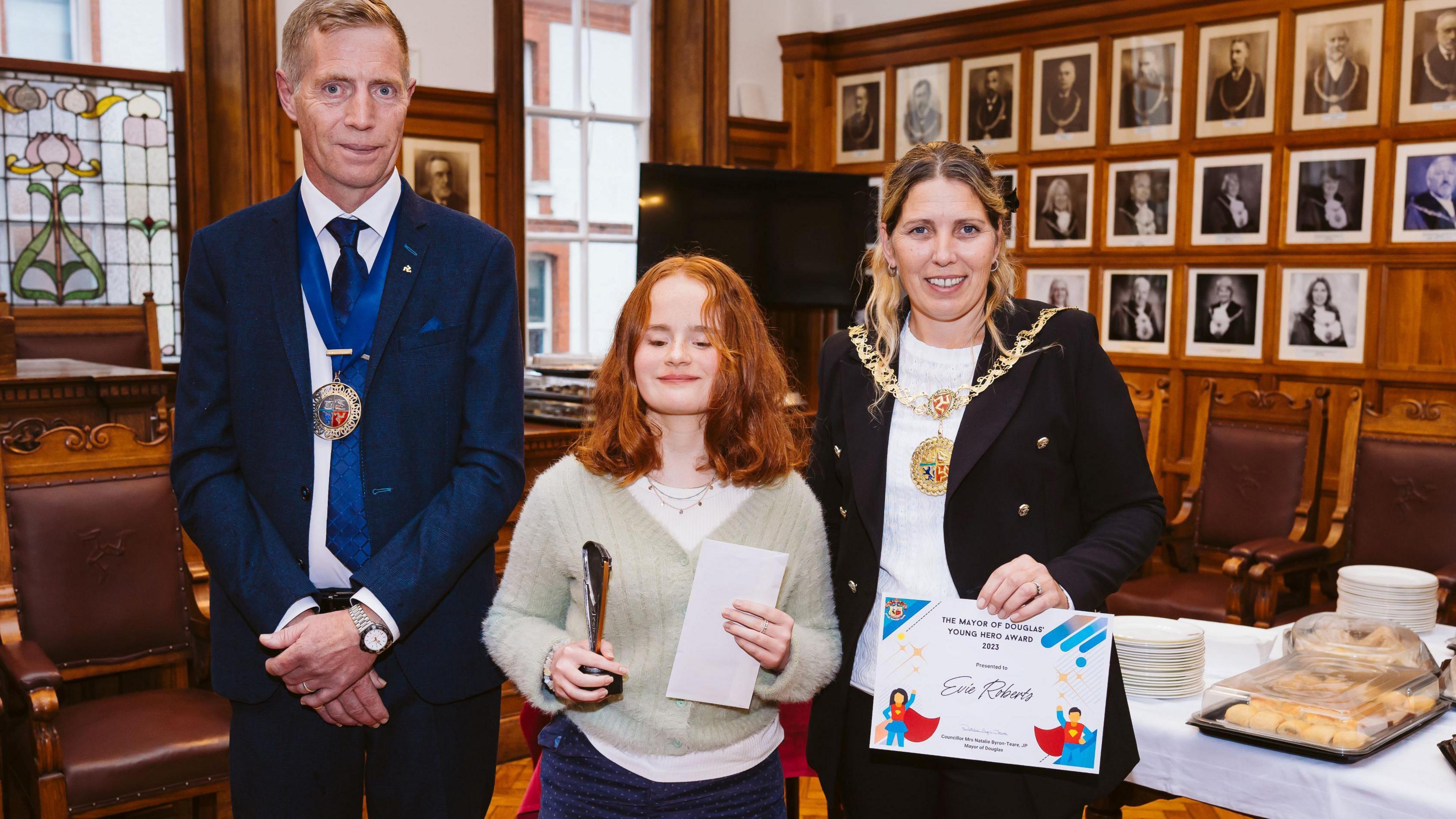 Evie, a teenage girl with short red hair, holds an award as she stands in the Douglas council chamber between Mayor Natalie Byron-Teare and consort Andy Teare.