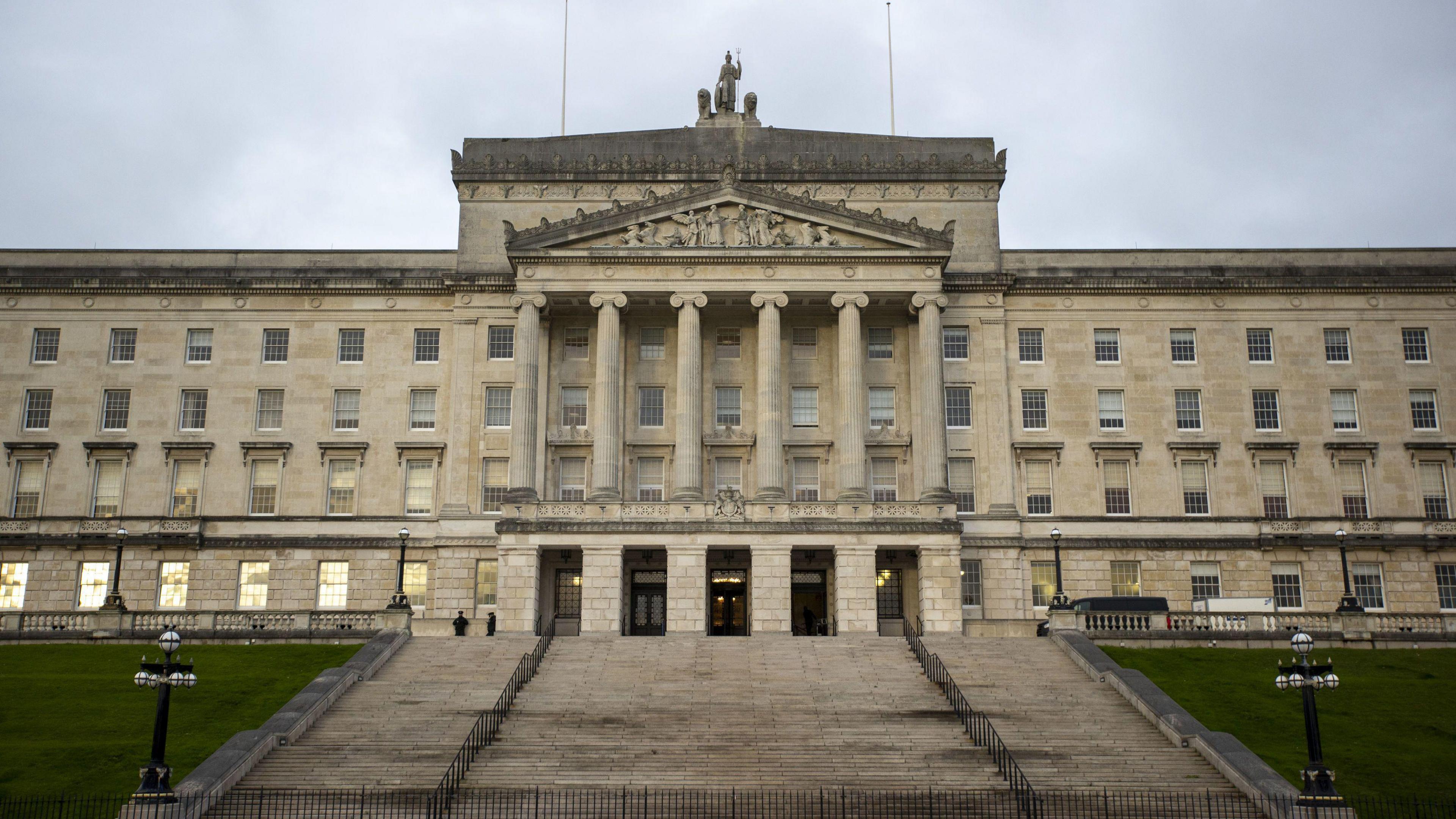 The steps leading up to the front of Stormont and the front of the building is visible. The building is a concrete colour and have six large central pillars. There are five doorways in the centre at the top of the steps. There is two black handrails on the steps. There is also visible grass on either side of the steps. 