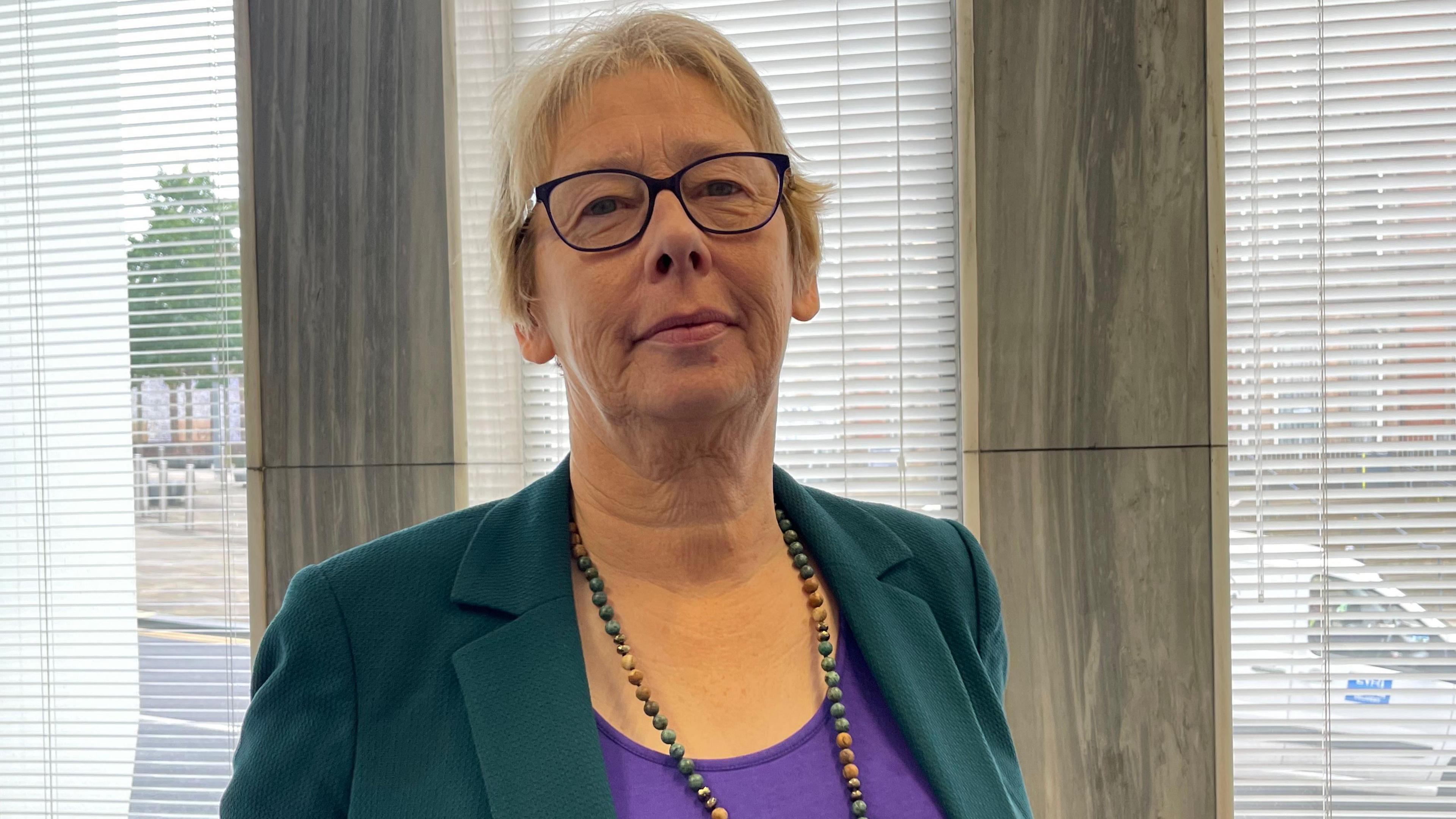 A woman with short blonde hair and wearing glasses, a green jacket, a purple top and a beaded necklace, stands next to an office window.