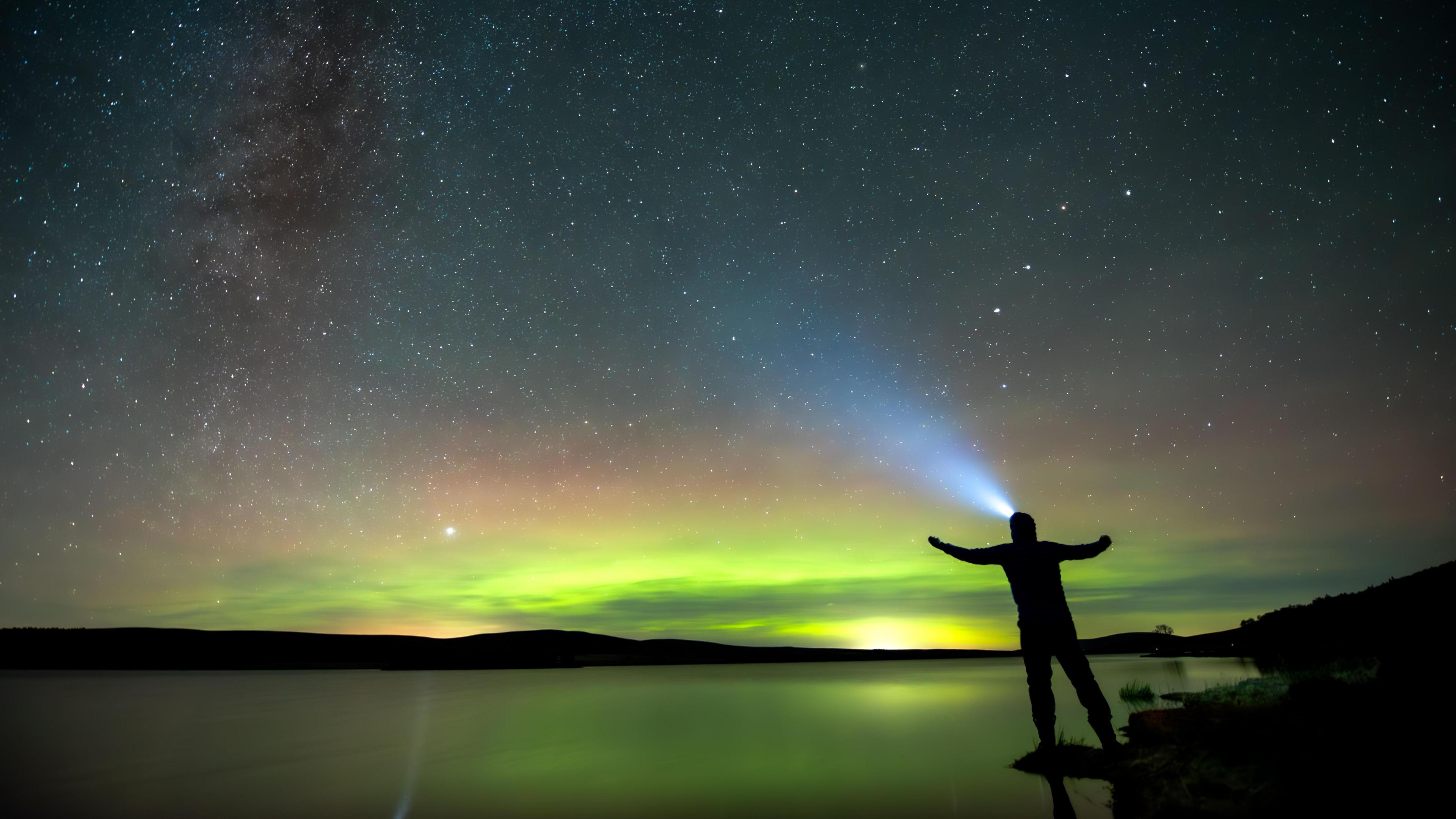 The silhouette of a man wearing a head torch in front of the green hues of the northern lights.