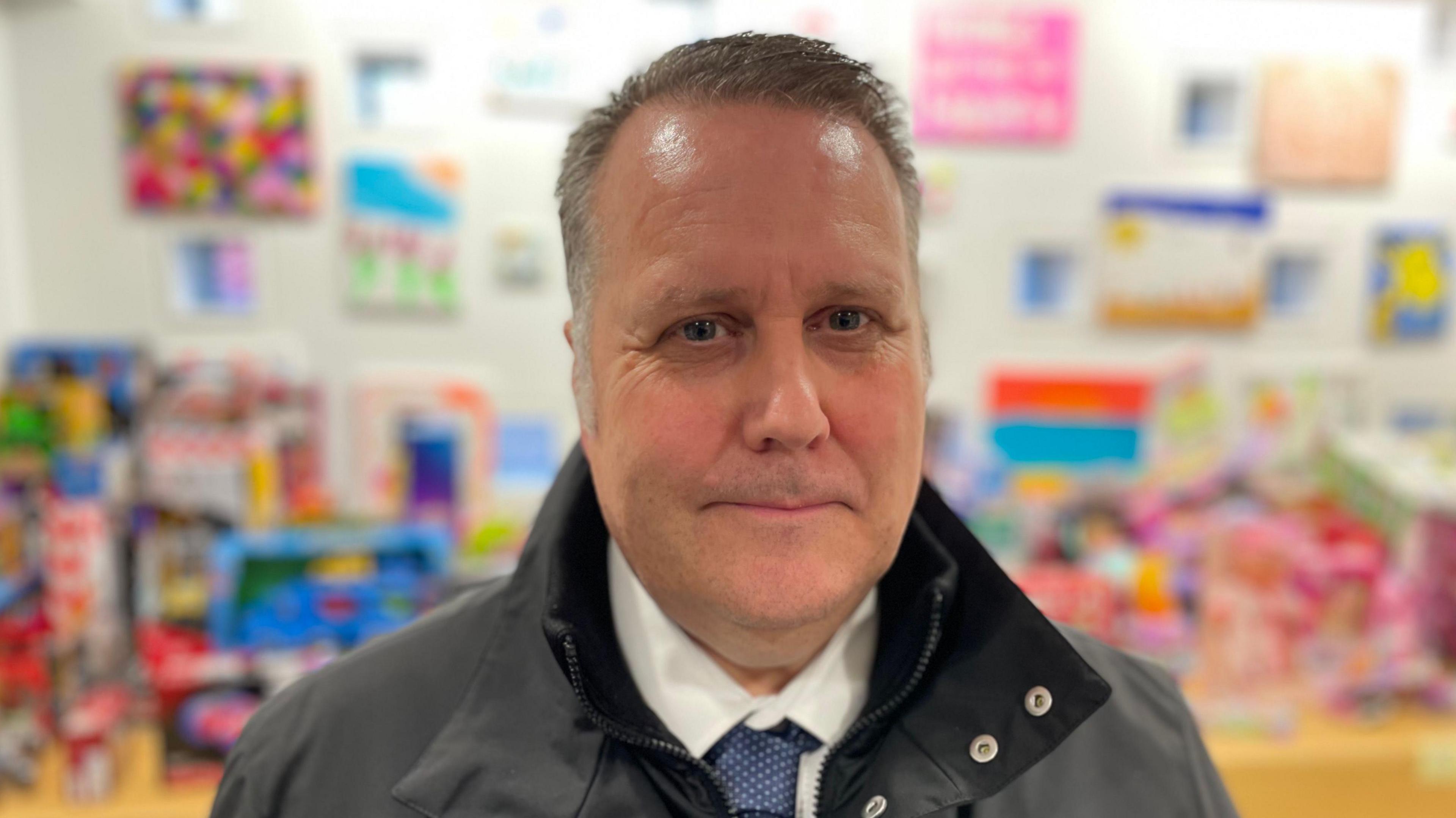 Mark Holloway looking at the camera. He has dark grey hair and is wearing a grey outdoor jacket, a white shirt and a blue tie with small white spots on it. There are blurred toys in the background.