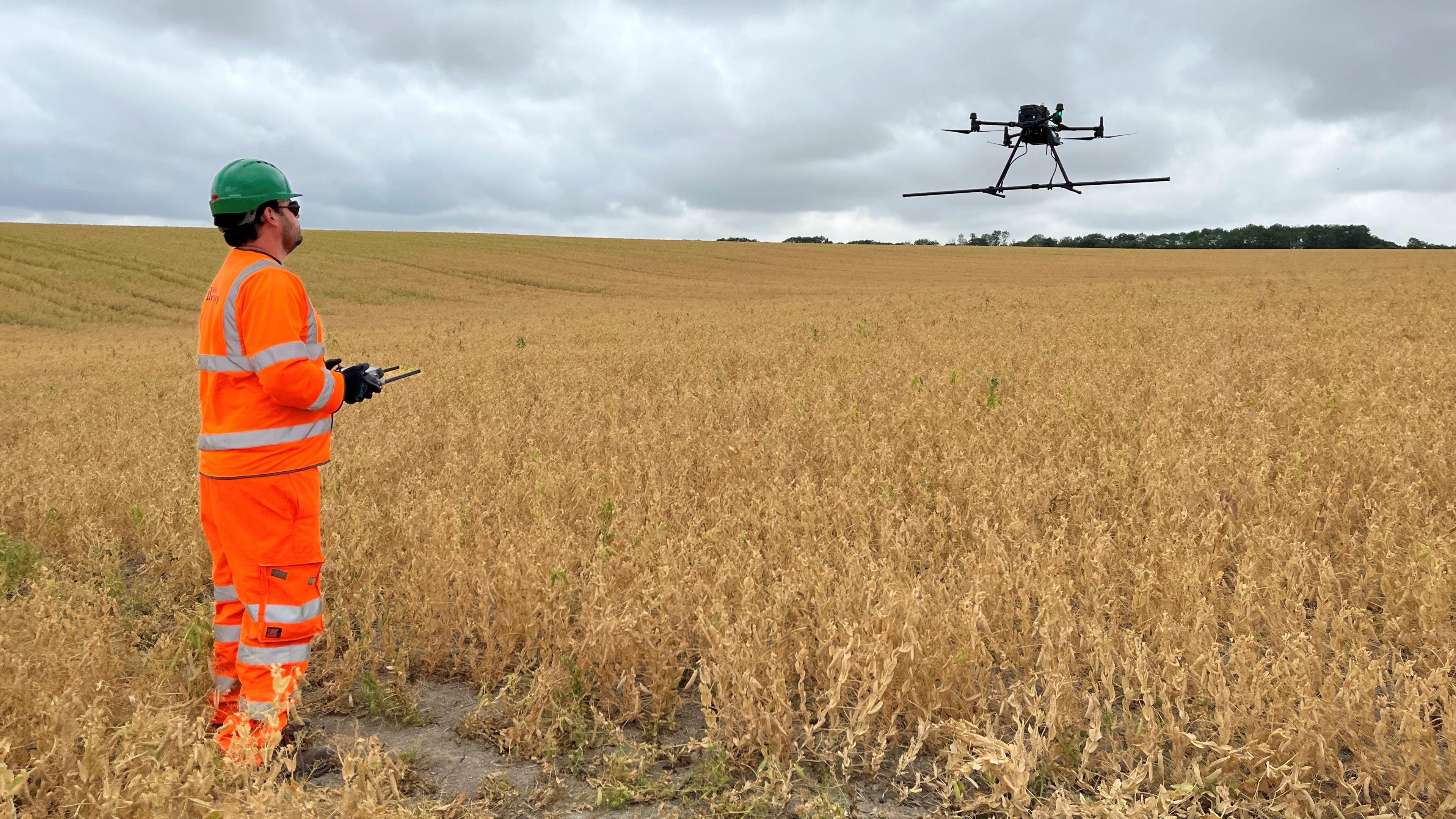 A man who is wearing an orange fluorescent protective clothing. He is also wearing a green hard hat and is flying a drone over a field.