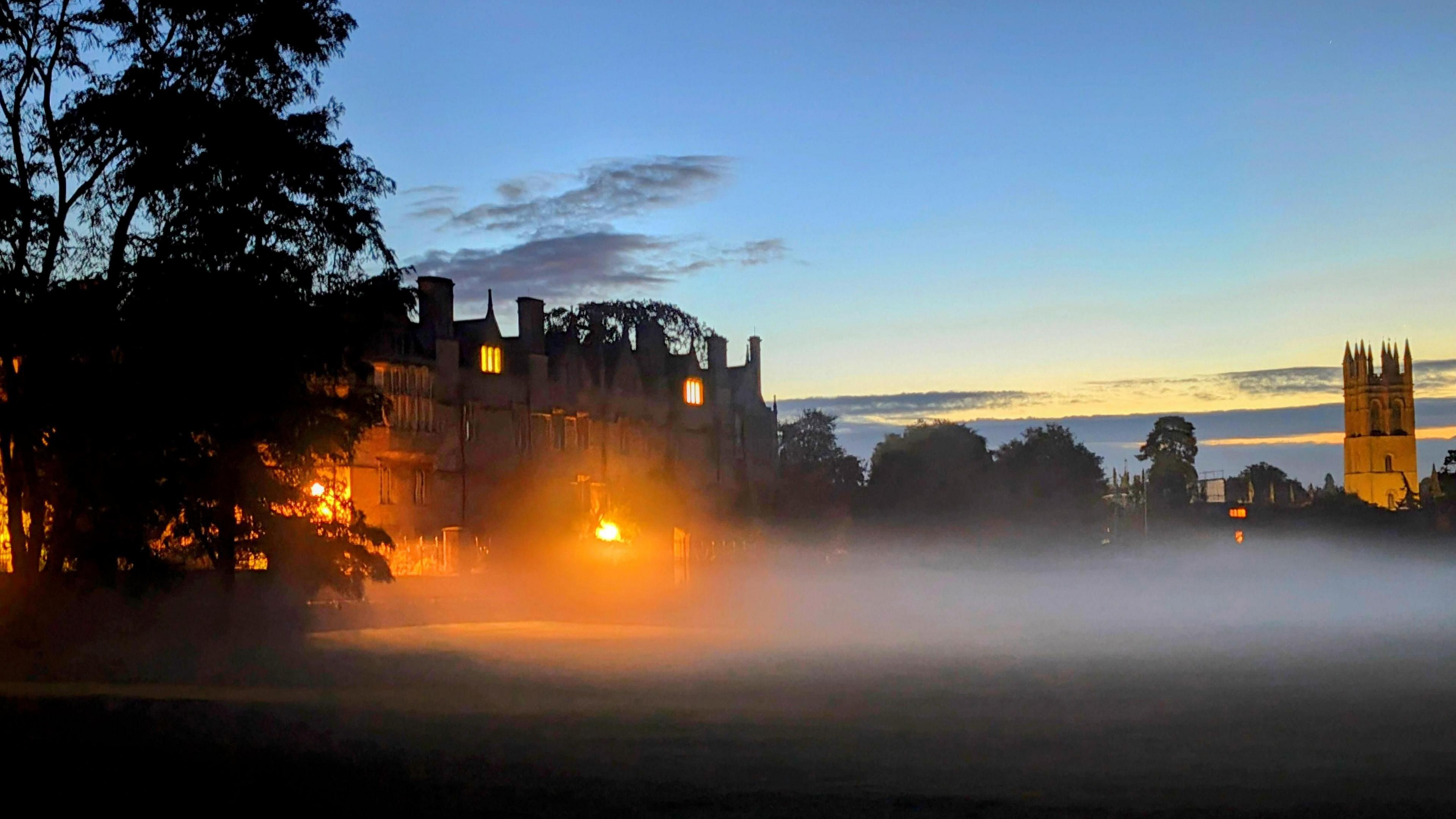 Mist appears over grass outside a large building in Oxford, with trees and a spire also seen in the background in low light.