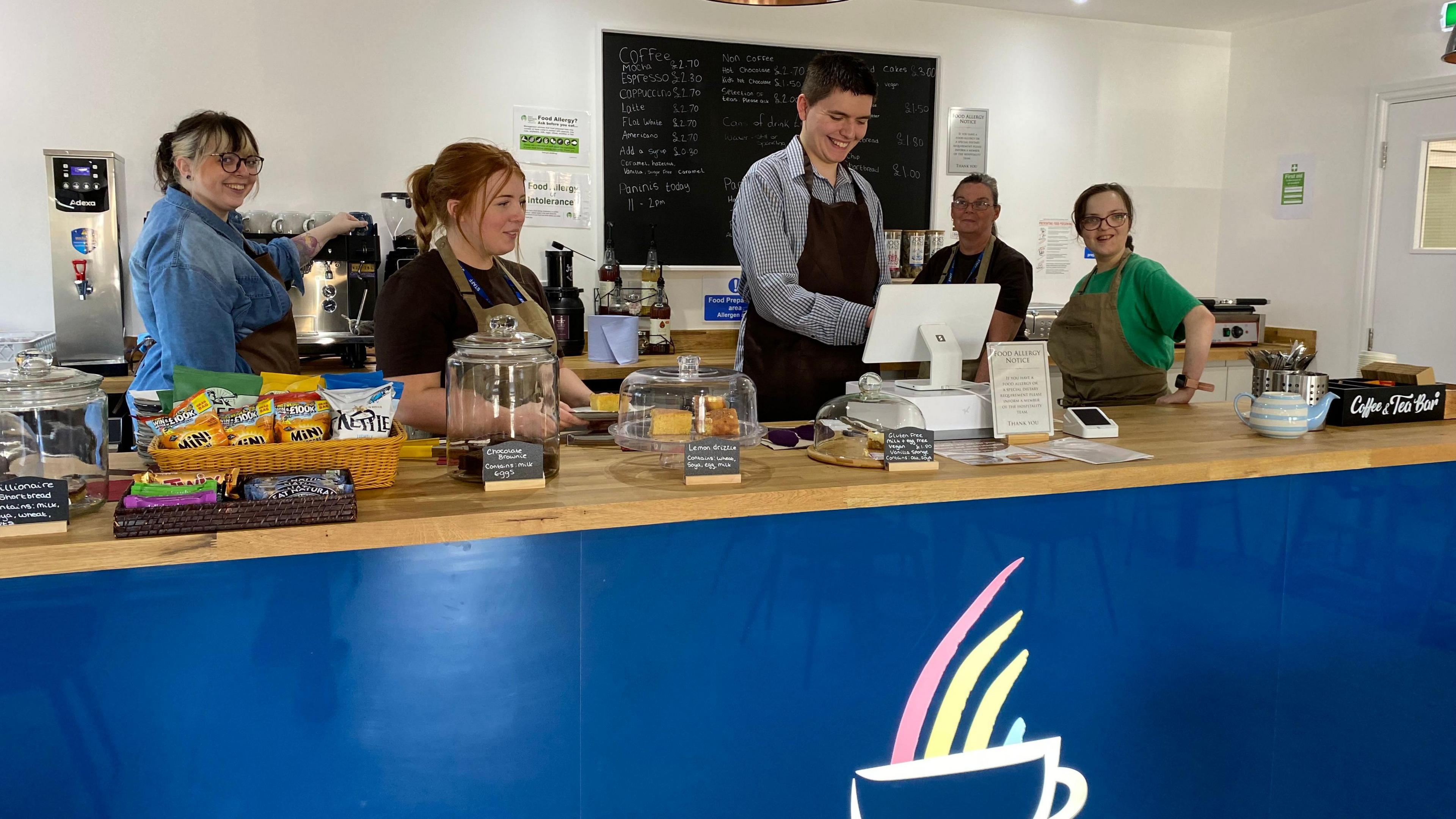 A large bar with crisps, chocolates and fresh cakes on display next to a cash register. Four young women and a man are standing behind the bar smiling and working in the cafe.