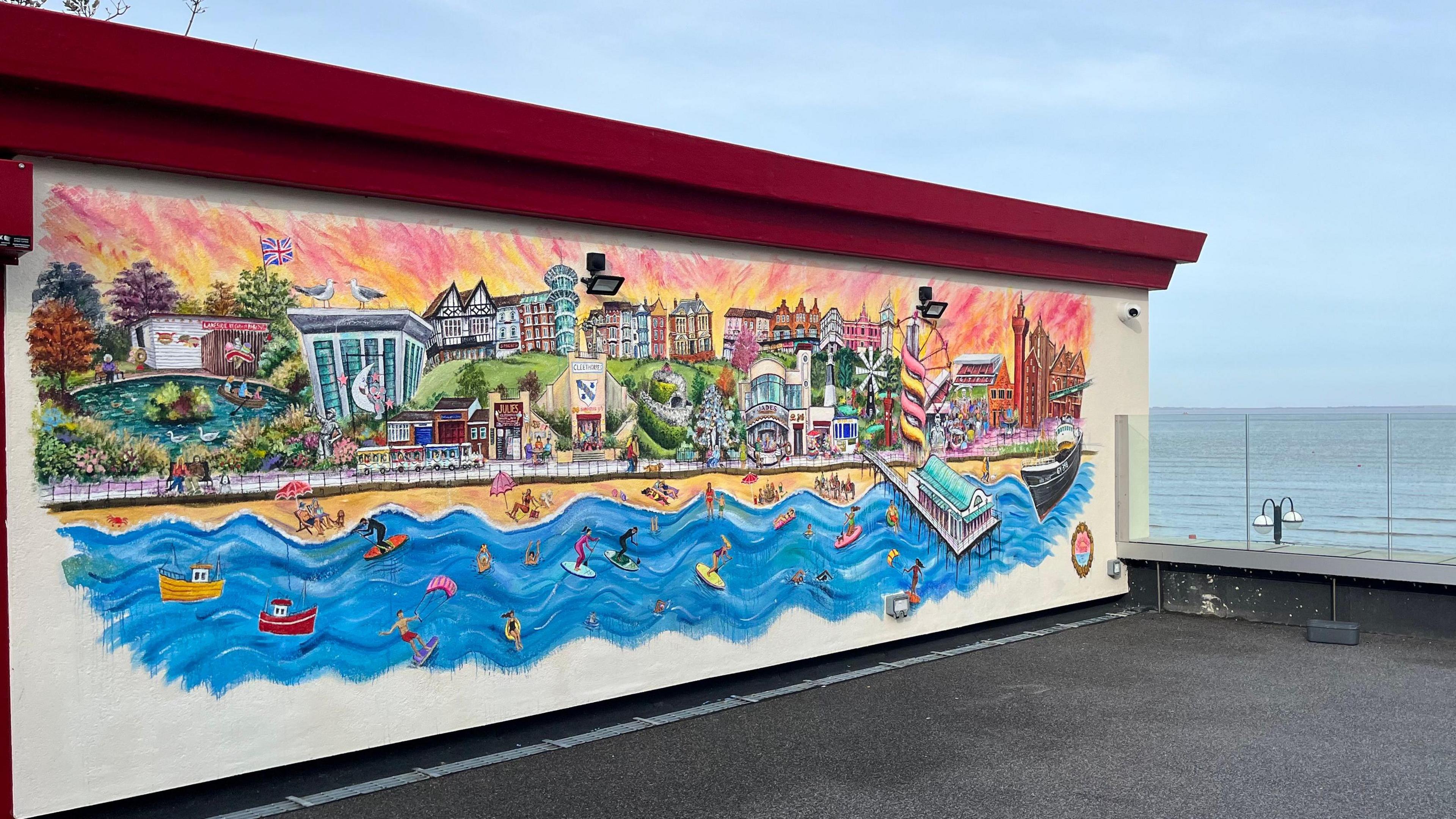 Brightly coloured mural painted on a wall, showing Cleethorpes beachfront, alongside beachgoers and surfers on the beach