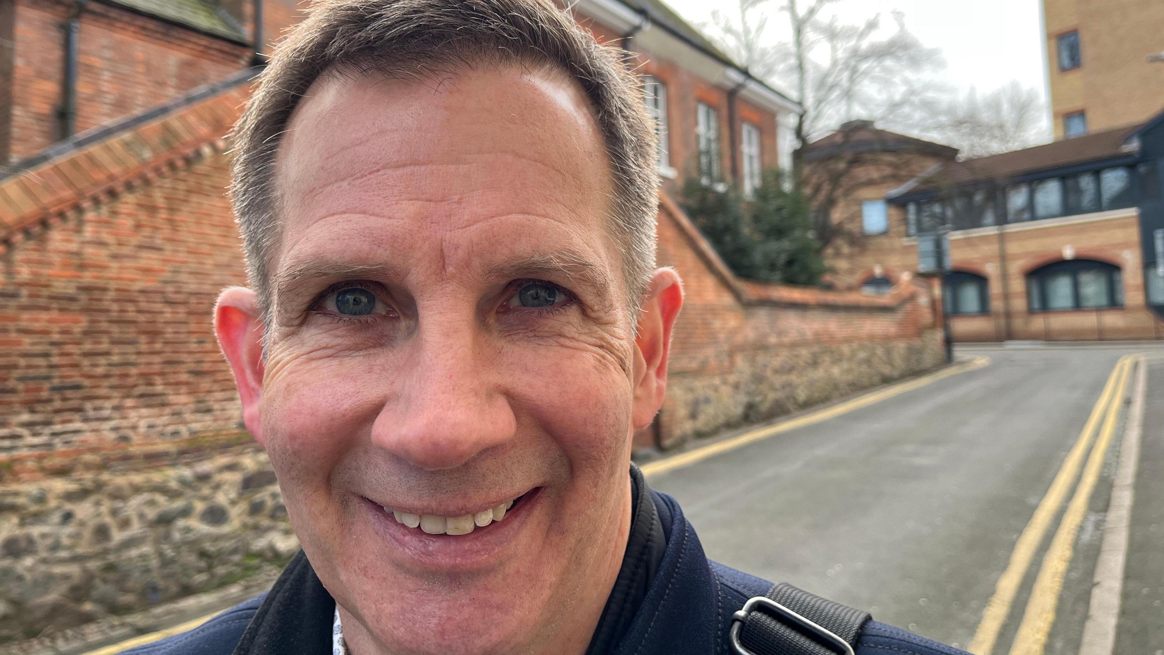 Nils smiles at a camera with a medieval wall to one side and a 1990s former tax office behind 