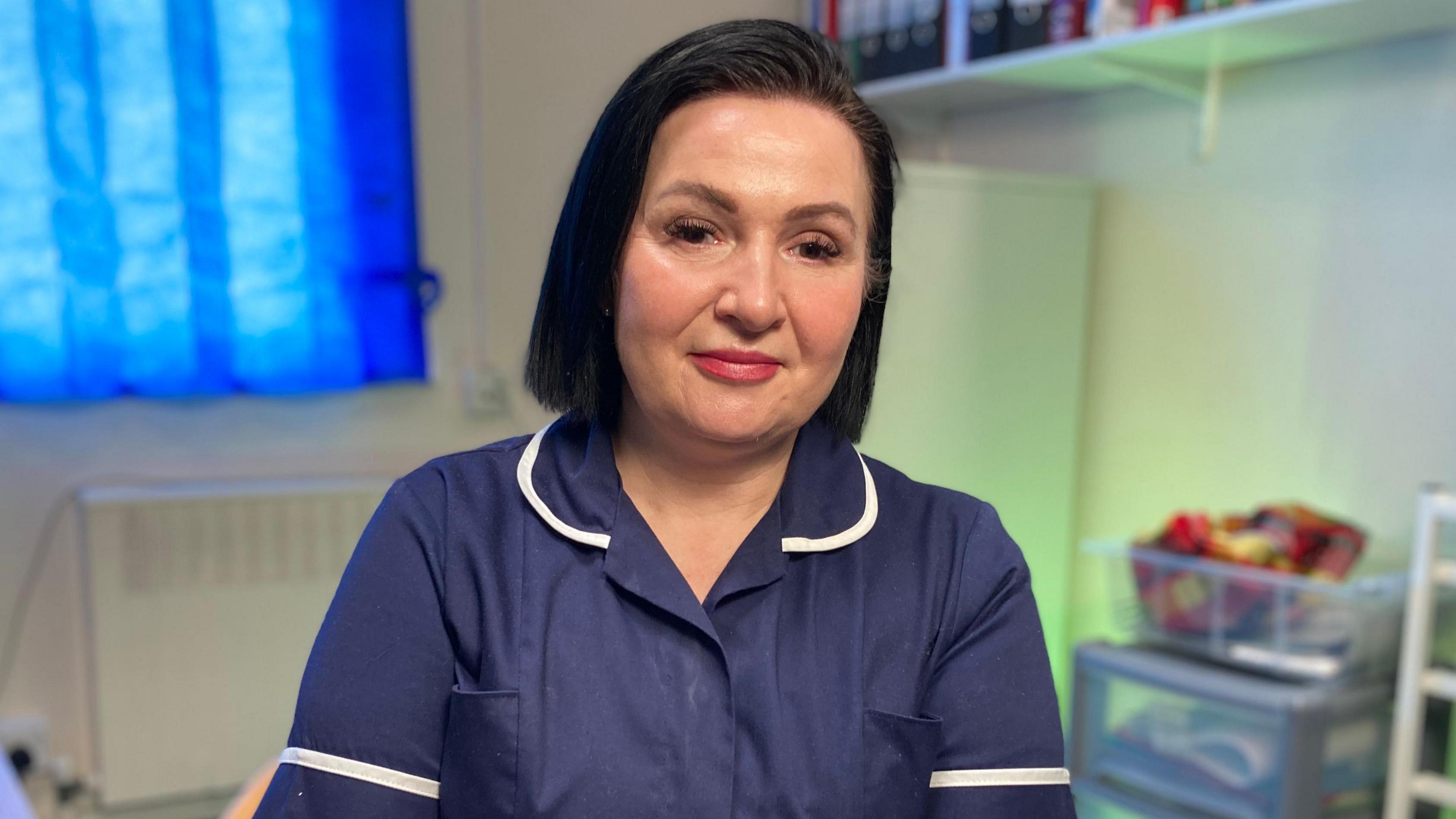 Kelly Harwood has black hair worn in a bob. She is wearing a navy blue nurse's uniform and is looking at the camera. There are medical supplies and folders on shelves behind her.