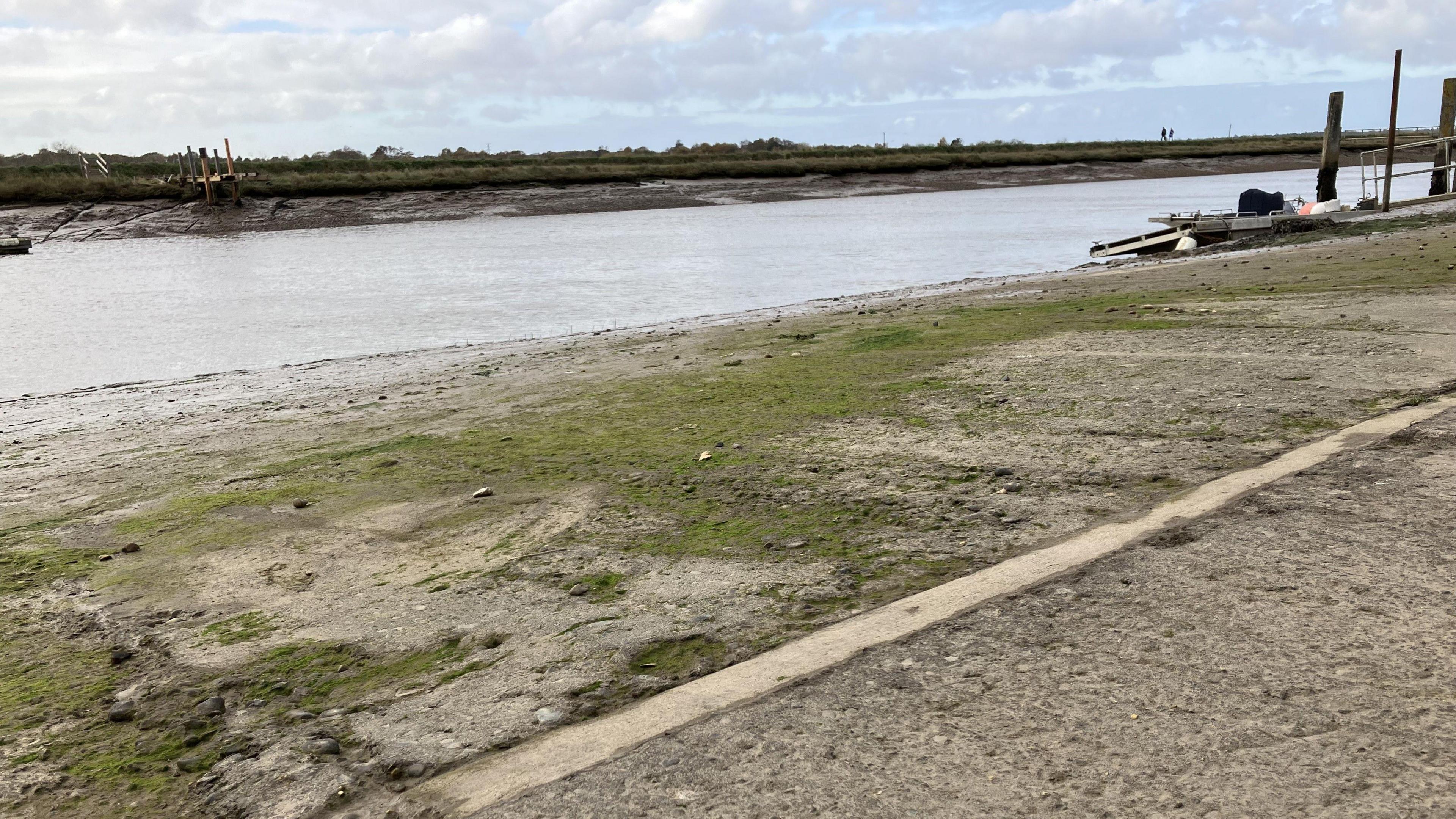 A grey and pitted concrete slipway with areas covered in green algae slopes towards the water of the River Blyth 