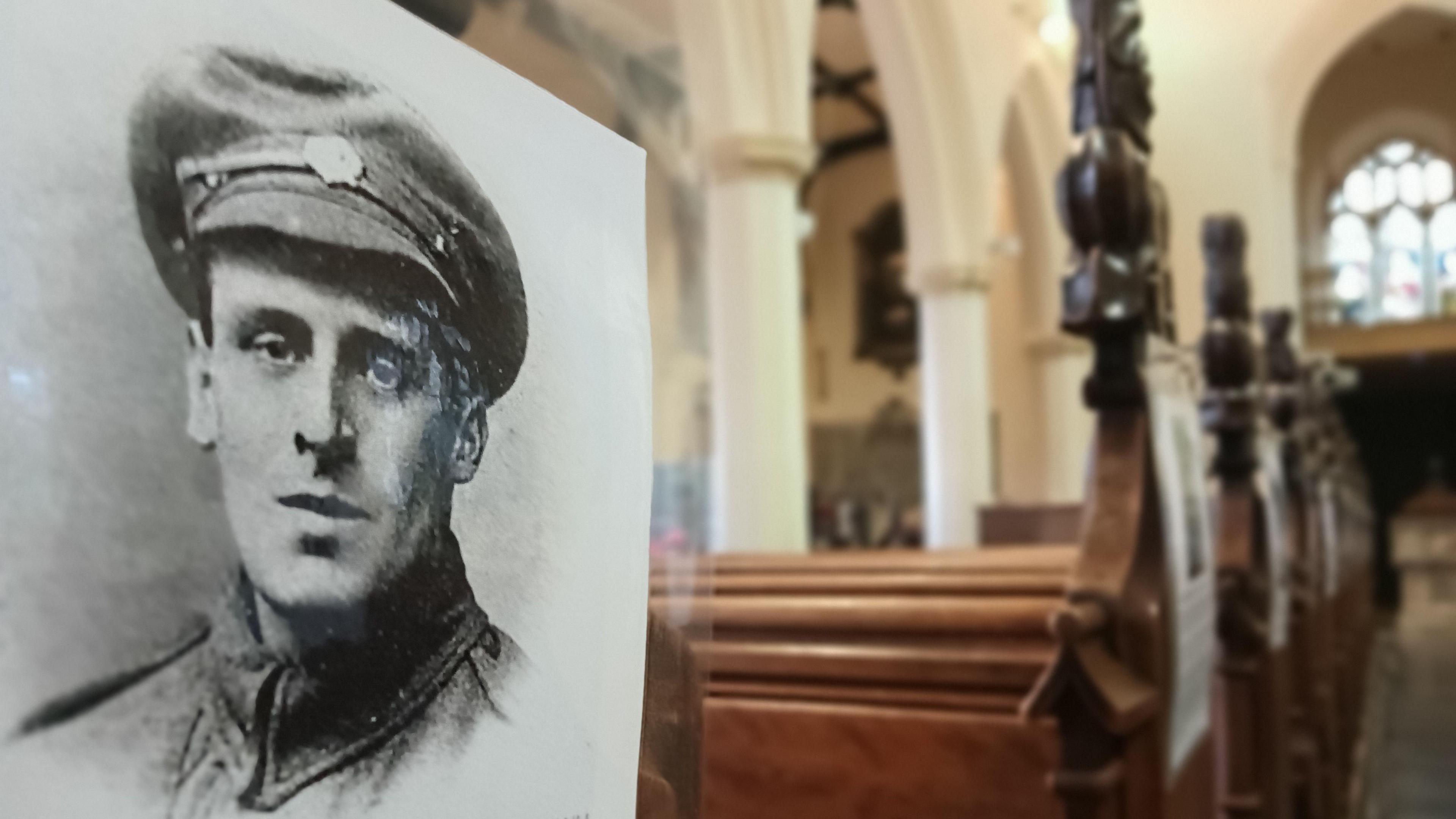 A close up of a portrait of a serviceman from World War One. It has been put in place at the end of a church pew. Further church pews can be seen behind it along with a stained glass window. 