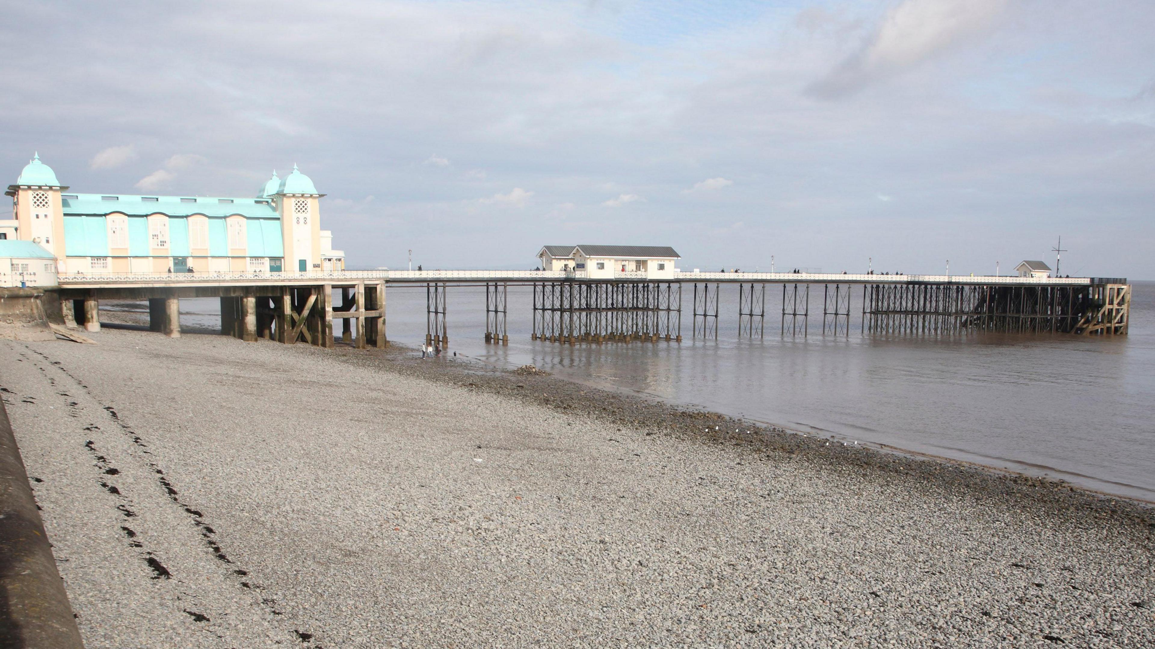 Penarth Pier and pavilion can be seen at the end of a stretch of pebbled beach. The pavilion is white with a blue roof, with domes on each end. The pier stretches out into the sea, with a white hut see in the middle of it.