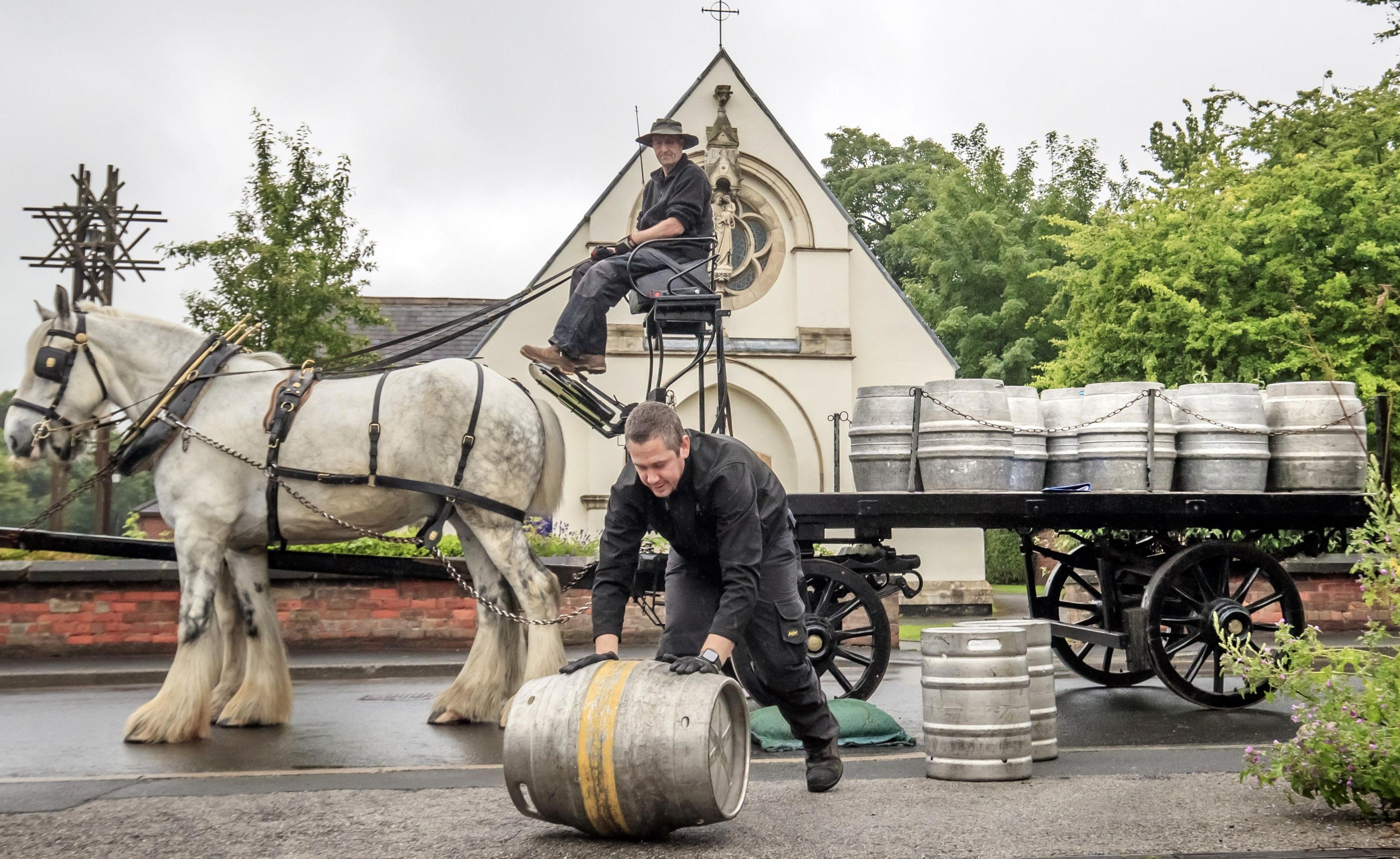 A man rolls a beer barrel as a man on a horse-drawn wagon, packed with more barrels, waits behind him.