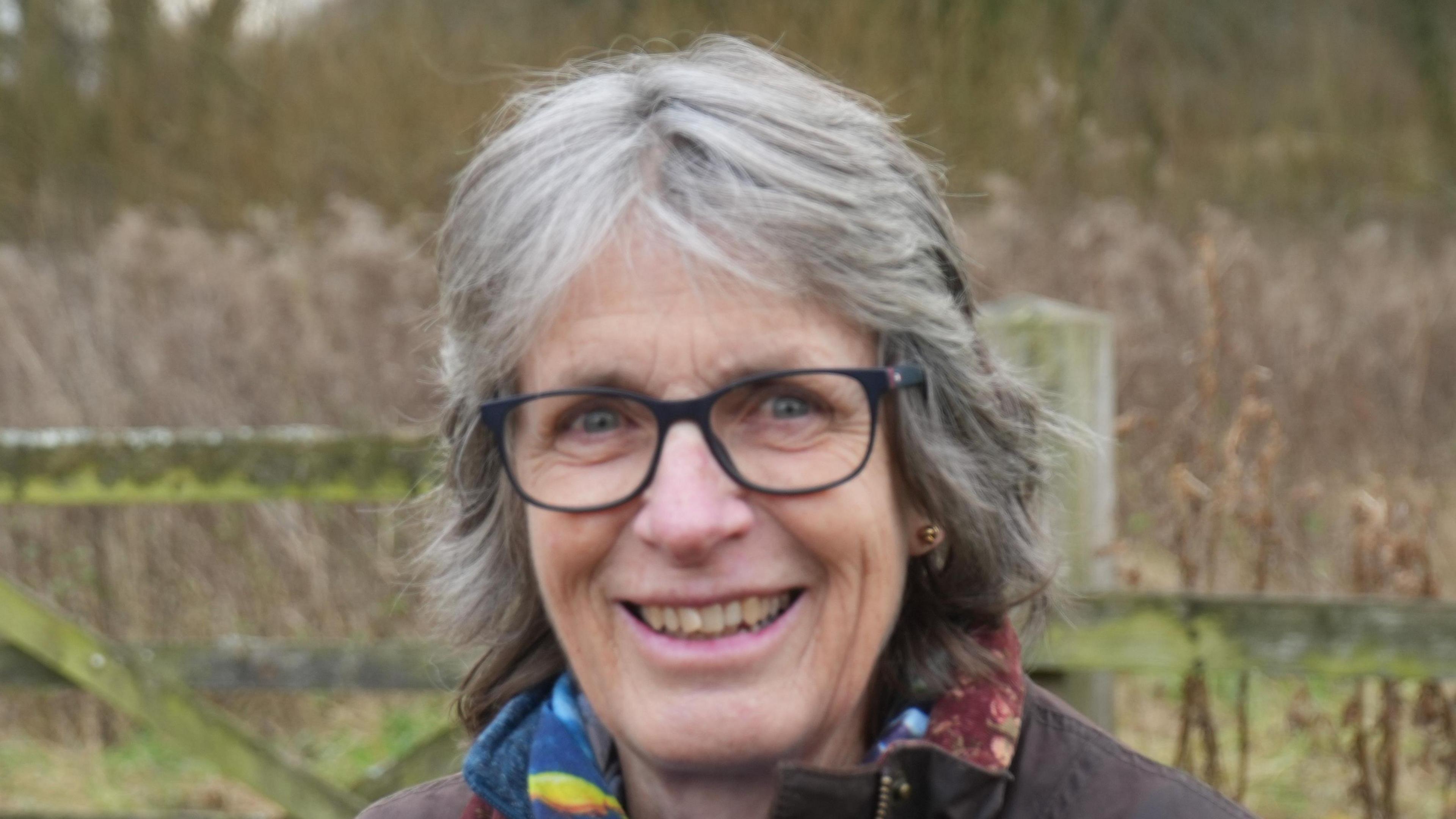 Penny Hemphill, who has grey and black short hair, smiles at the camera while she stands in a field. She is wearing black glasses, a brown coat and a multi-coloured scarf. 