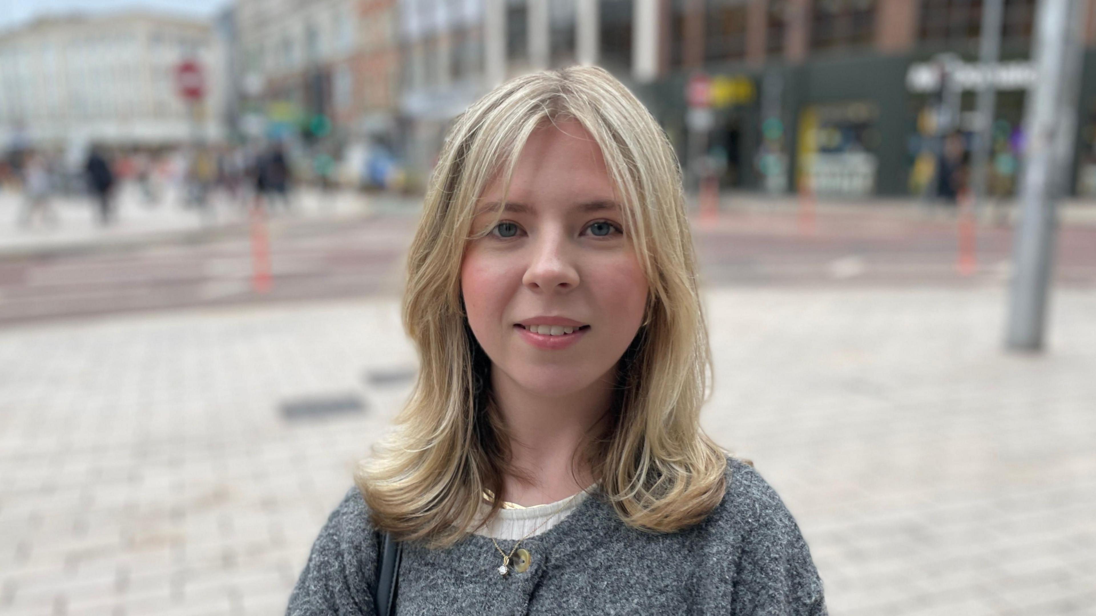 Ellie Ferguson is wearing a grey cardigan with a white top underneath. She is also wearing a silver chain. She has blonde hair. The background is blurred. 