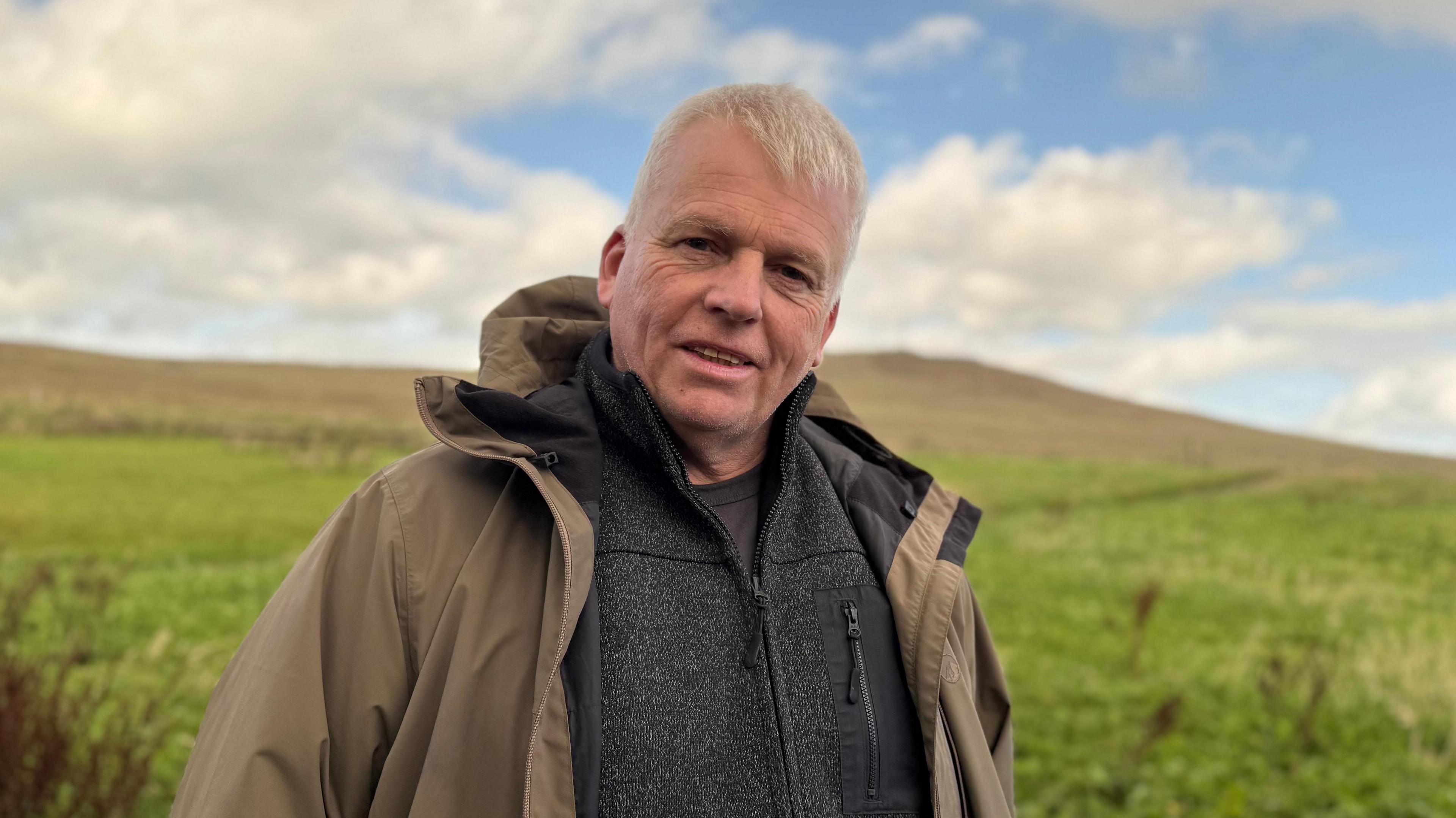 Peter Harper, a man with short, white hair, looks at the camera while standing in the Belfast Hills on a sunny day. He is wearing a dark t-shirt and fleece over a brown hooded coat.