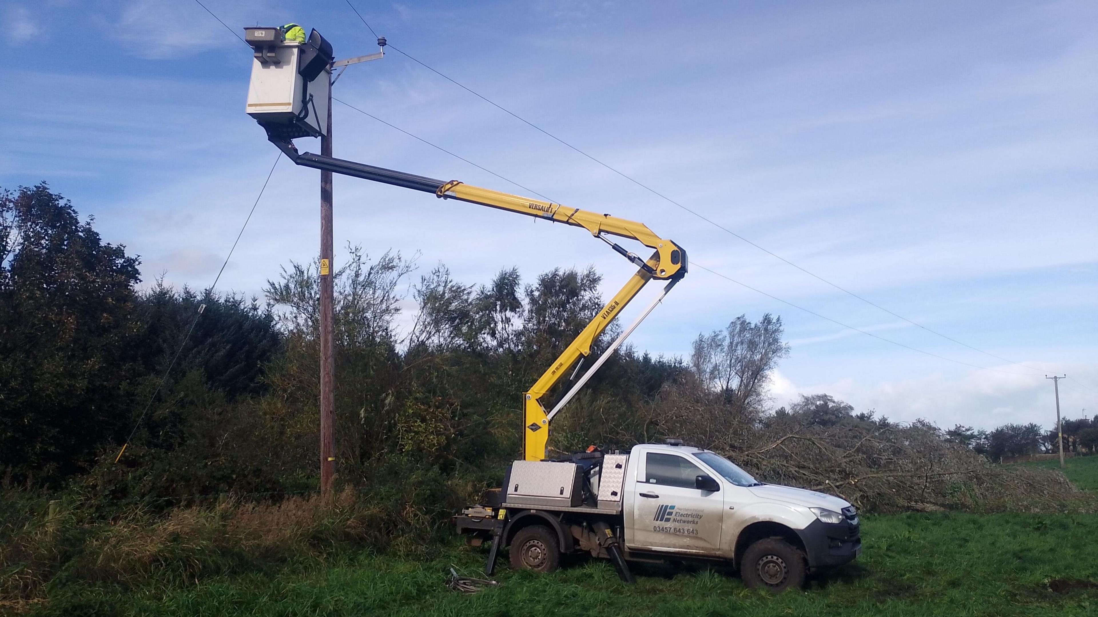 A NIE Networks truck is parked beside a power line pole as a small cherrypicker attached to the back hoists a worker into the air to fix a broken powerline.