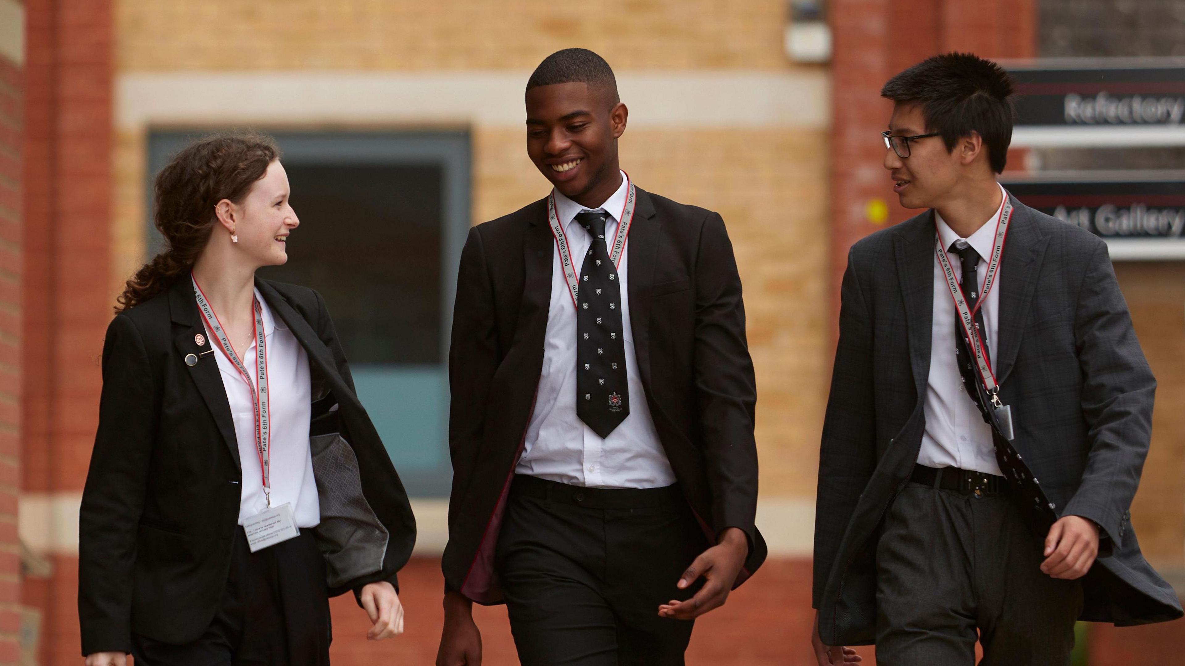 Three students are walking on the Pate's Grammar School campus, one girl and two boys about the age of 15 to 17 years-old. They are all wearing white shirts, and dark coloured suits. The two boys are wearing black ties with the Pate's Grammar logo printed on them. They all have white and red lanyards around their necks. 