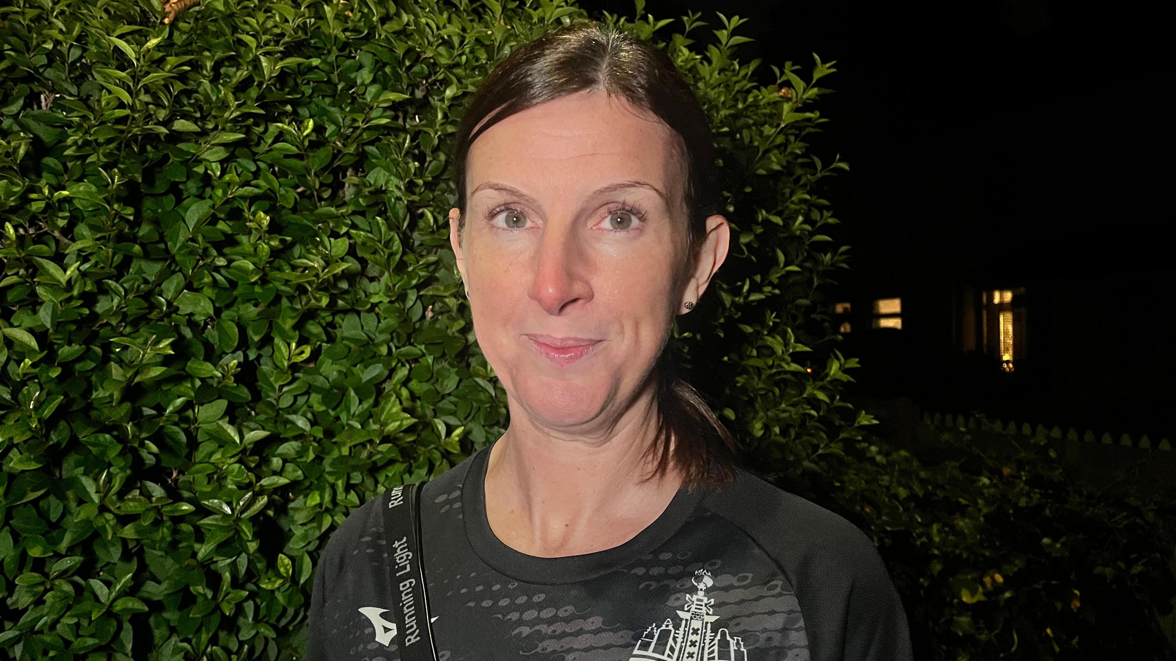 Runner Jeannine Ursell is wearing a black sport shirt. She has dark hair, tied back. She is standing in front of a green shrub under a night sky.