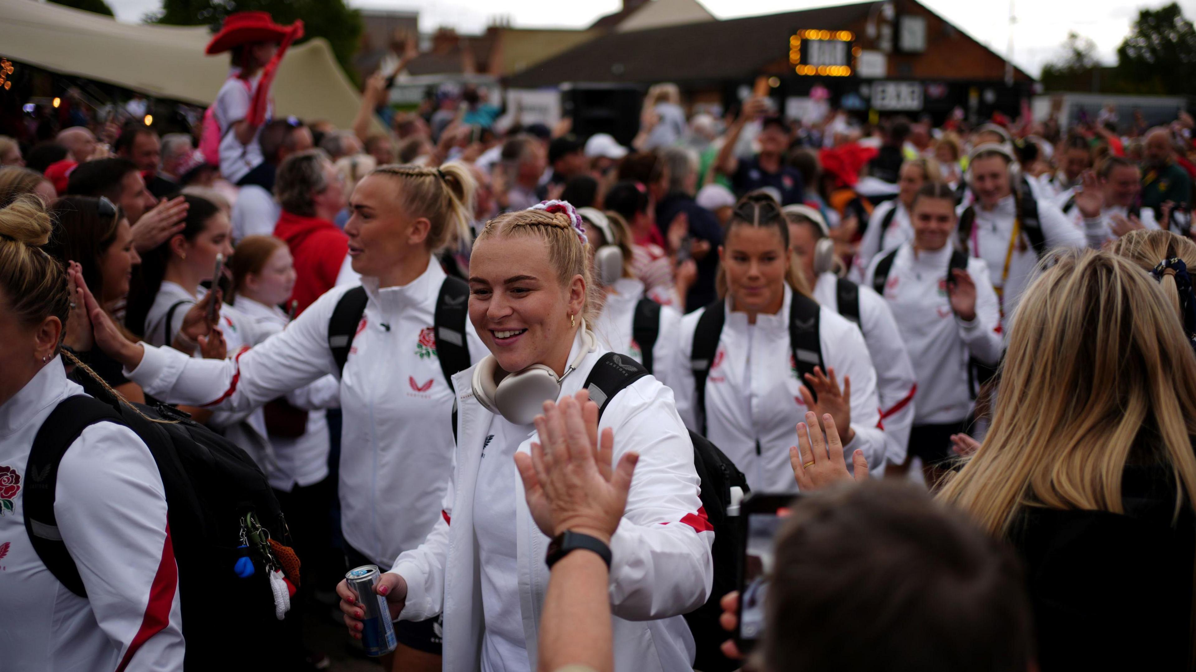 The England team arrives before the Women's Rugby World Cup 2025 Pool A match at Franklin's Gardens, Northampton. The team are wearing white track suits, and are high-fiving members of the crowd. 