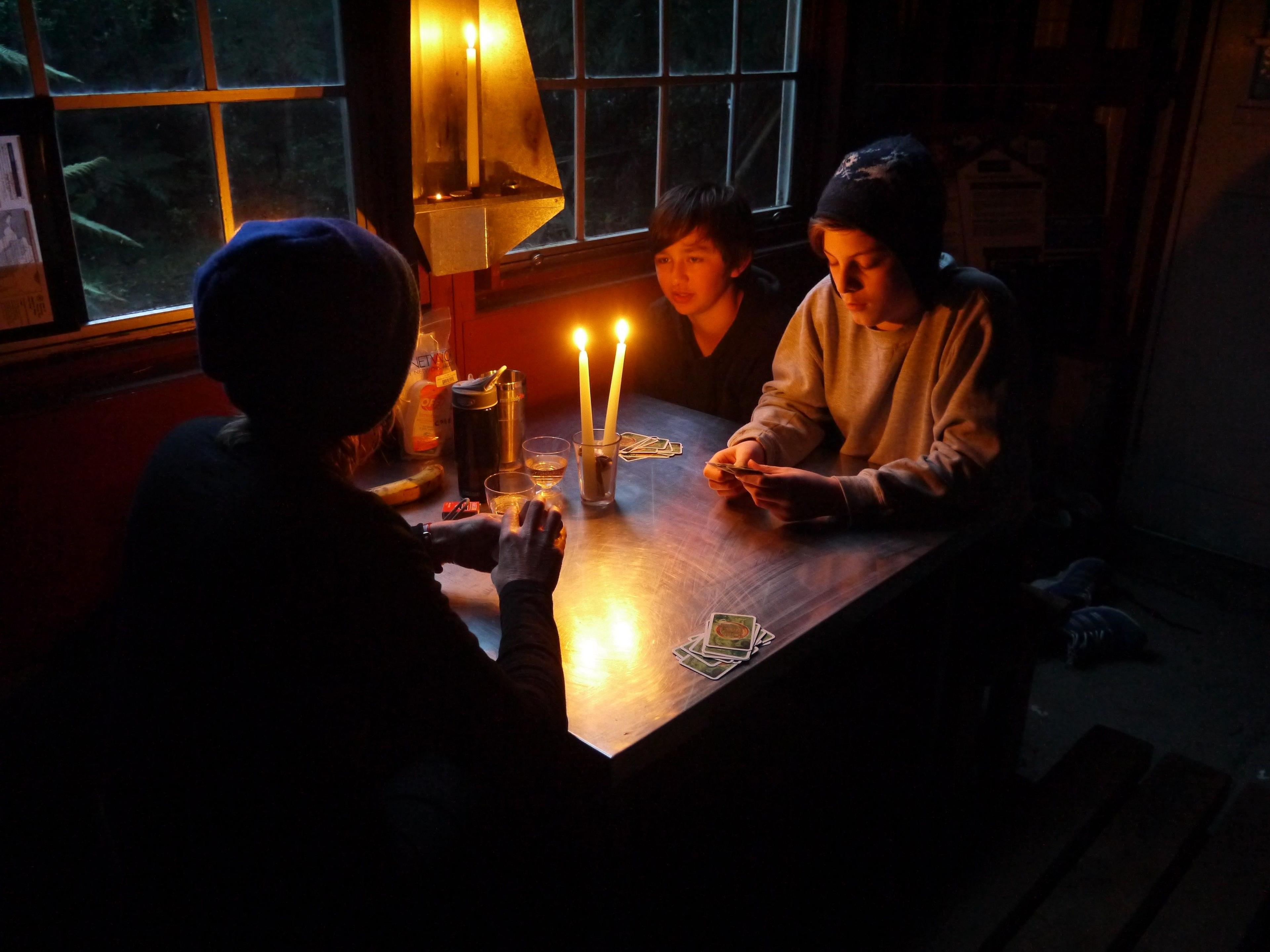 Three people playing cards at a table by candlelight. They are sitting beside a window and it is dark outside.