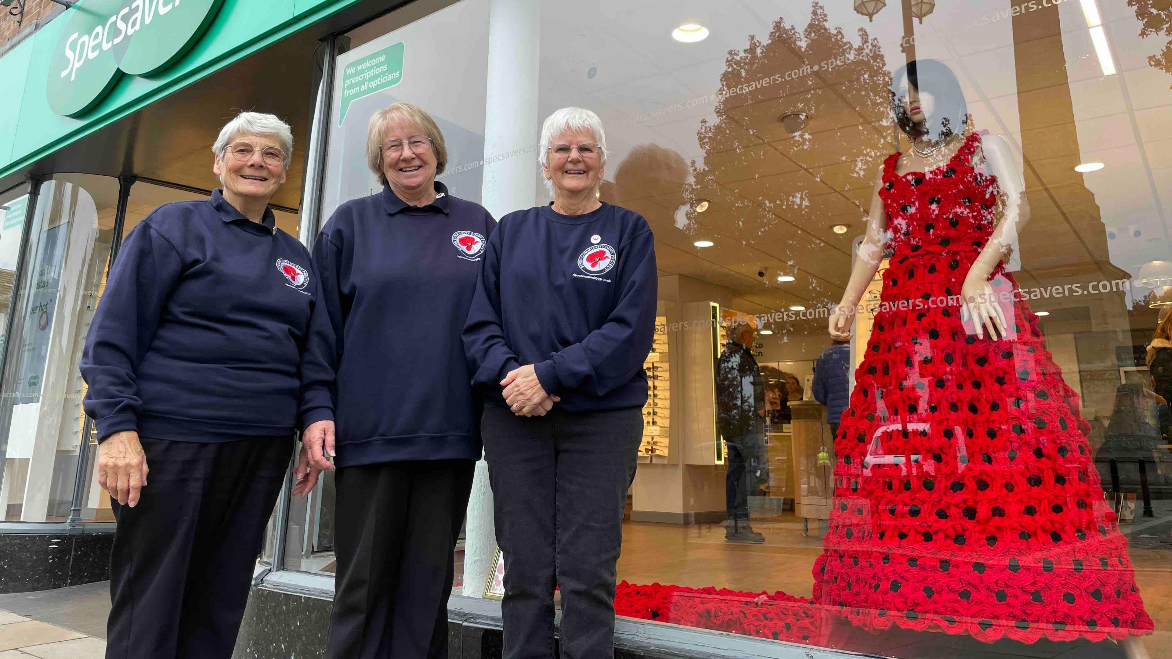 Three older women stand outside a Specsavers shop, wearing matching navy sweatshirts with a red poppy logo. Behind them, a mannequin in the store window displays a red dress made entirely of knitted poppies.
