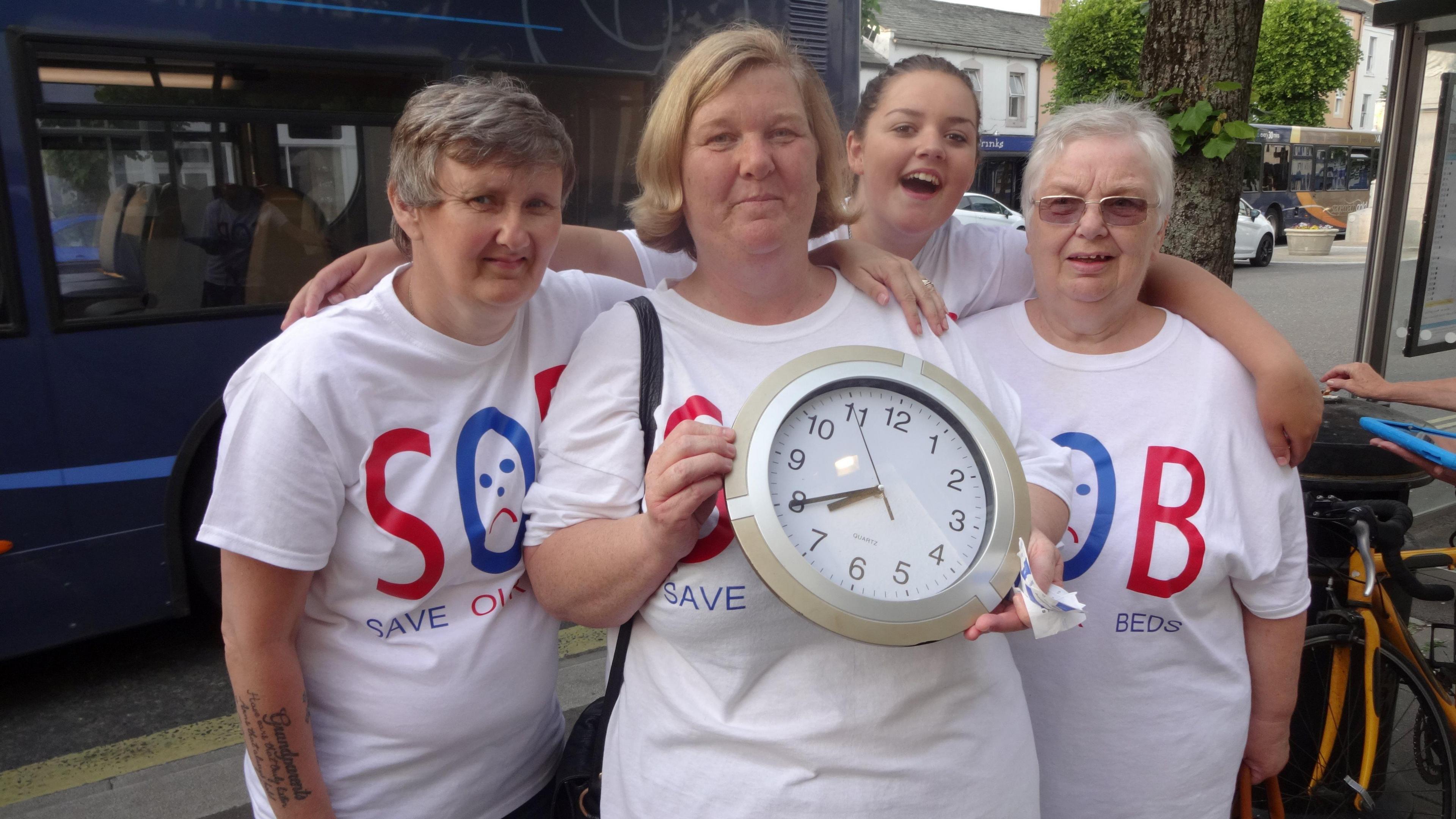 Three women and a girl wearing SOB (Save our Beds) t-shirts and holding a clock. They are pictured at a bus stop in Cockermouth.