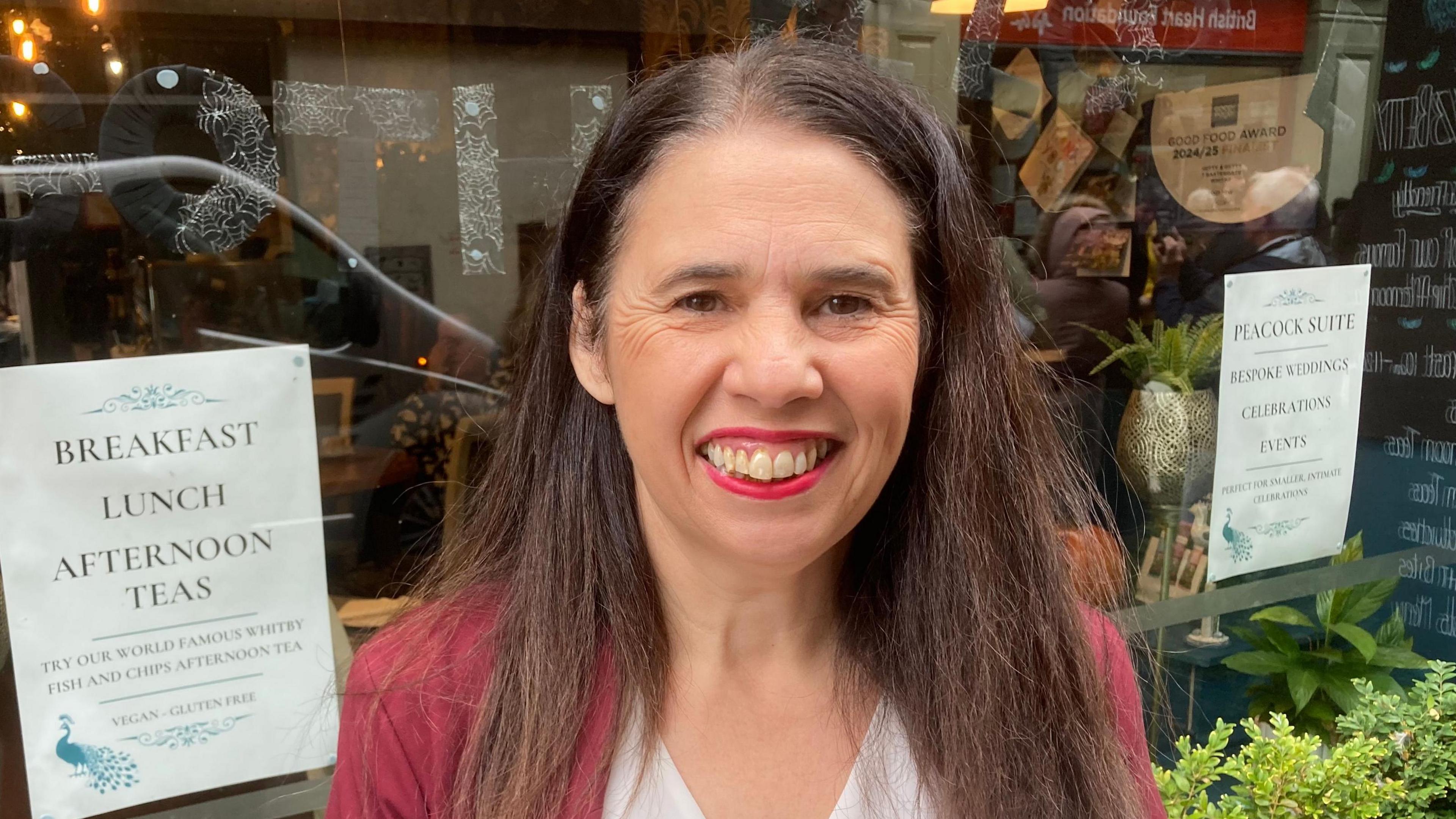 A woman with long brown hair smiles widely at the camera. She is stood in front of a shop or restaurant window. She wears a maroon cardigan and pink lipstick.