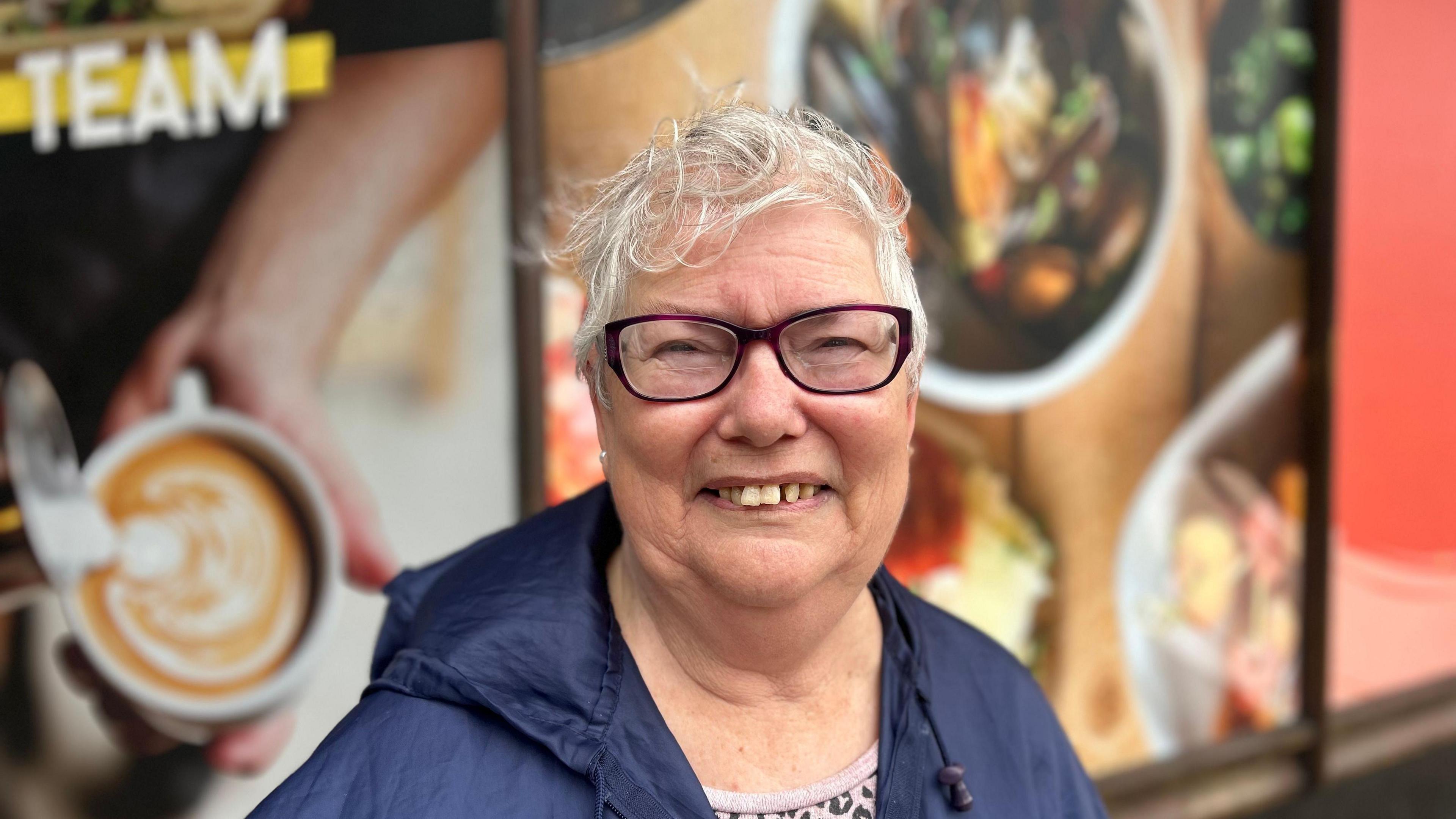 A lady smiling outside of the Hammonds building, she has a blue coat and glasses on, her eyes are squinted and you can see her teeth within her grin.