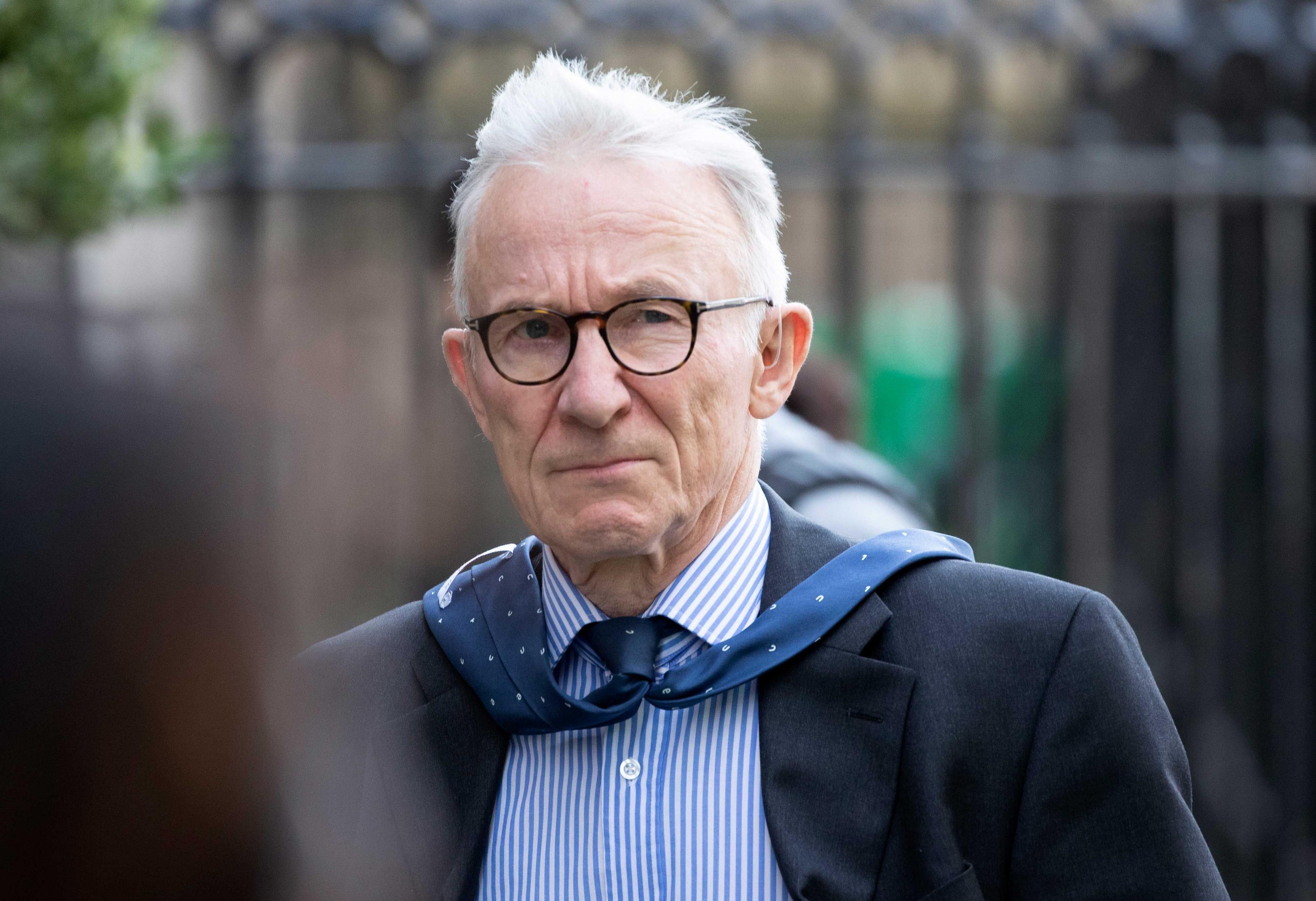 Lord Brodie walking on a street with railings behind him. He is wearing a dark blue suit, striped shirt and glasses. The wind has blown his tie over his shoulders.