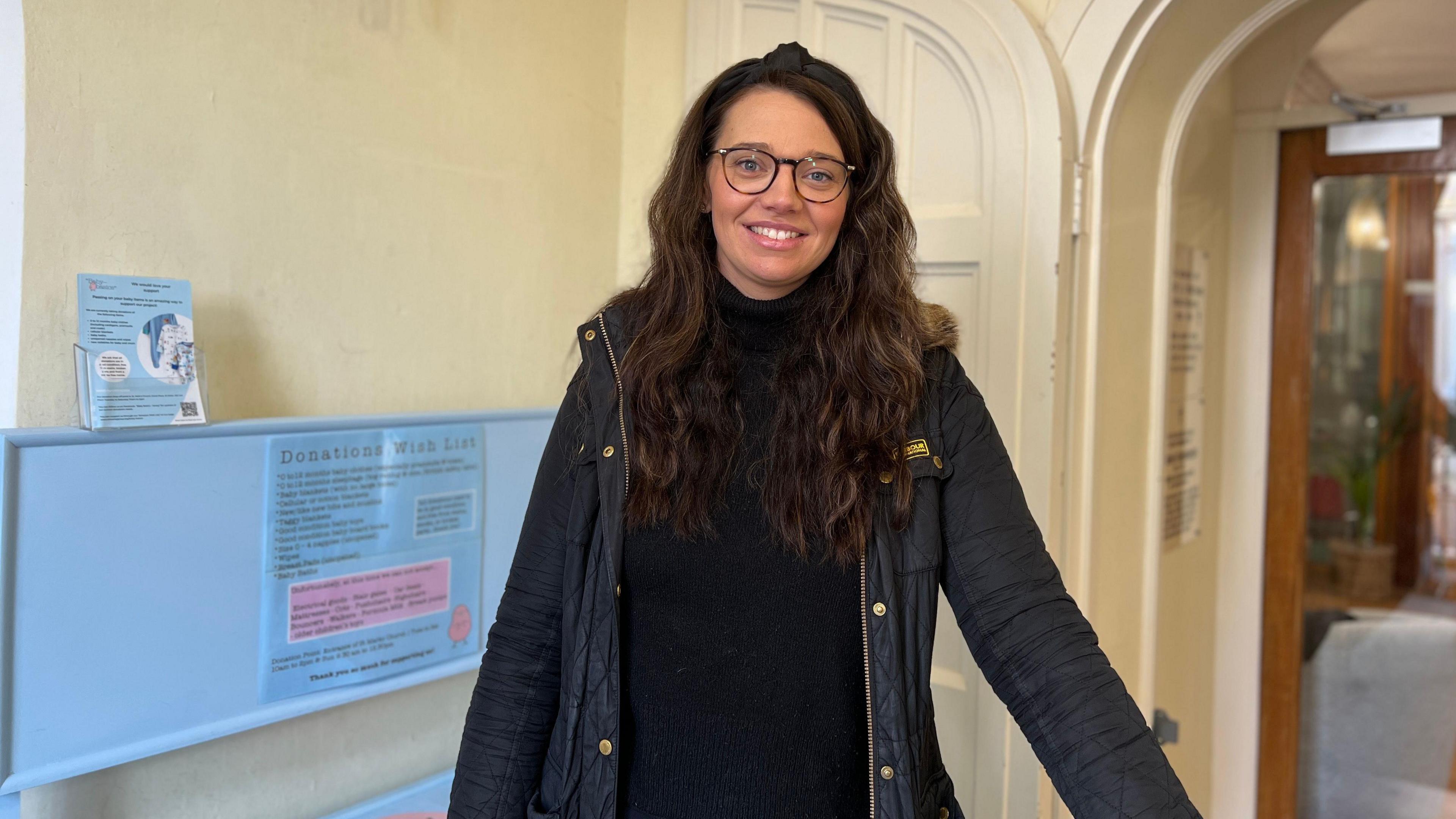 Stephanie smiles at the camera as the donation box is to her left. She is wearing a big black coat with a black jumper under it. She has long dark brown hair which comes below her shoulders. She is wearing glasses with a black rim. 