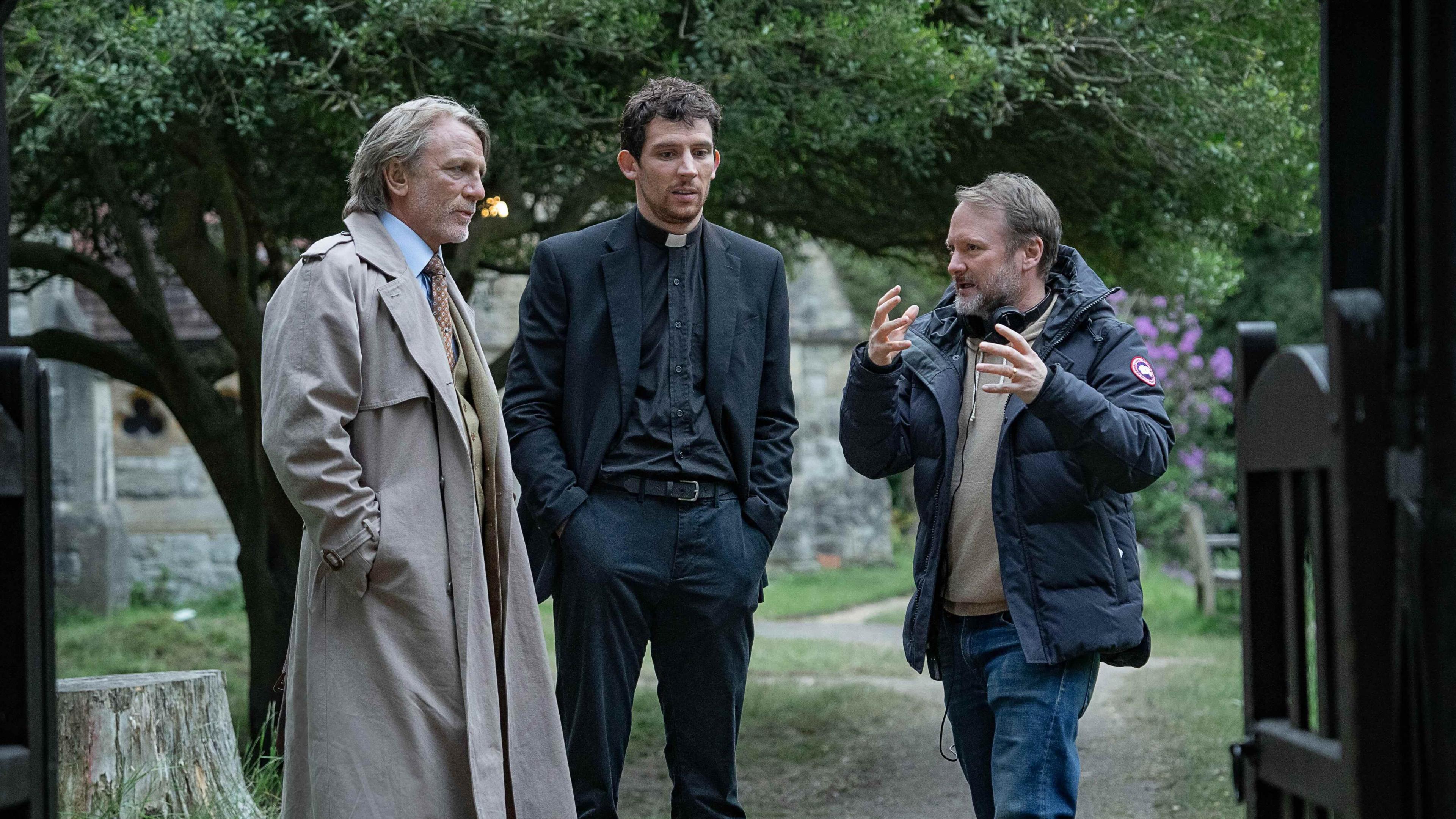 (Left to right) Daniel Craig and Josh O'Connor and writer-director Rian Johnson are standing just beyond the entrance to a church graveyard with the church itself in the background. Johnson, on the right, is gesturing to Craig and O'Connor, who are listening intently. Craig is wearing a long beige raincoat with shirt and tie, while O'Connor is dressed as a clergyman in dog collar and black shirt, jacket and trousers. 