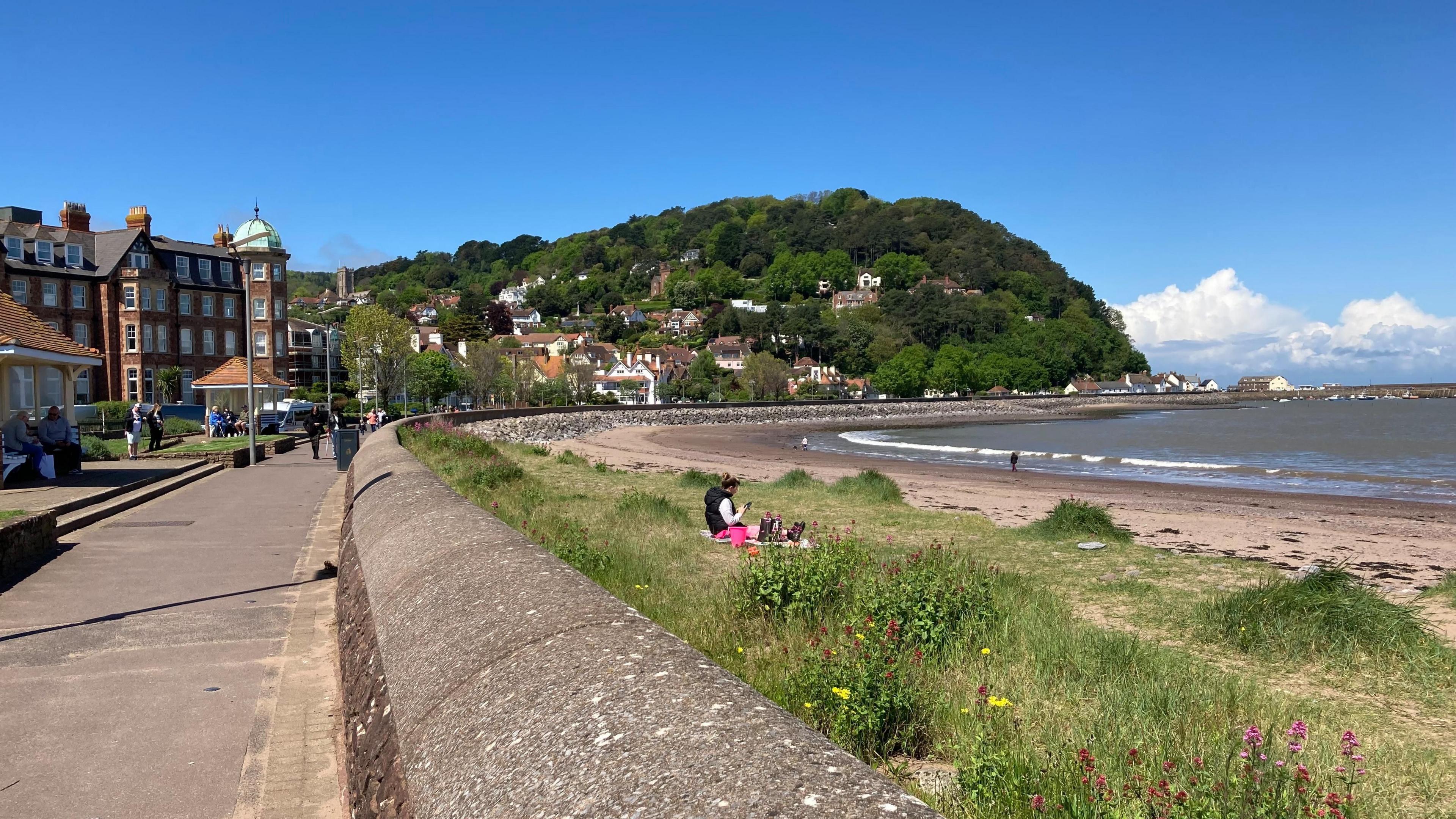 The seafront at Minehead with the Esplanade running behind sand dunes and the beach on a sunny day with the town centre and cliffs in the background