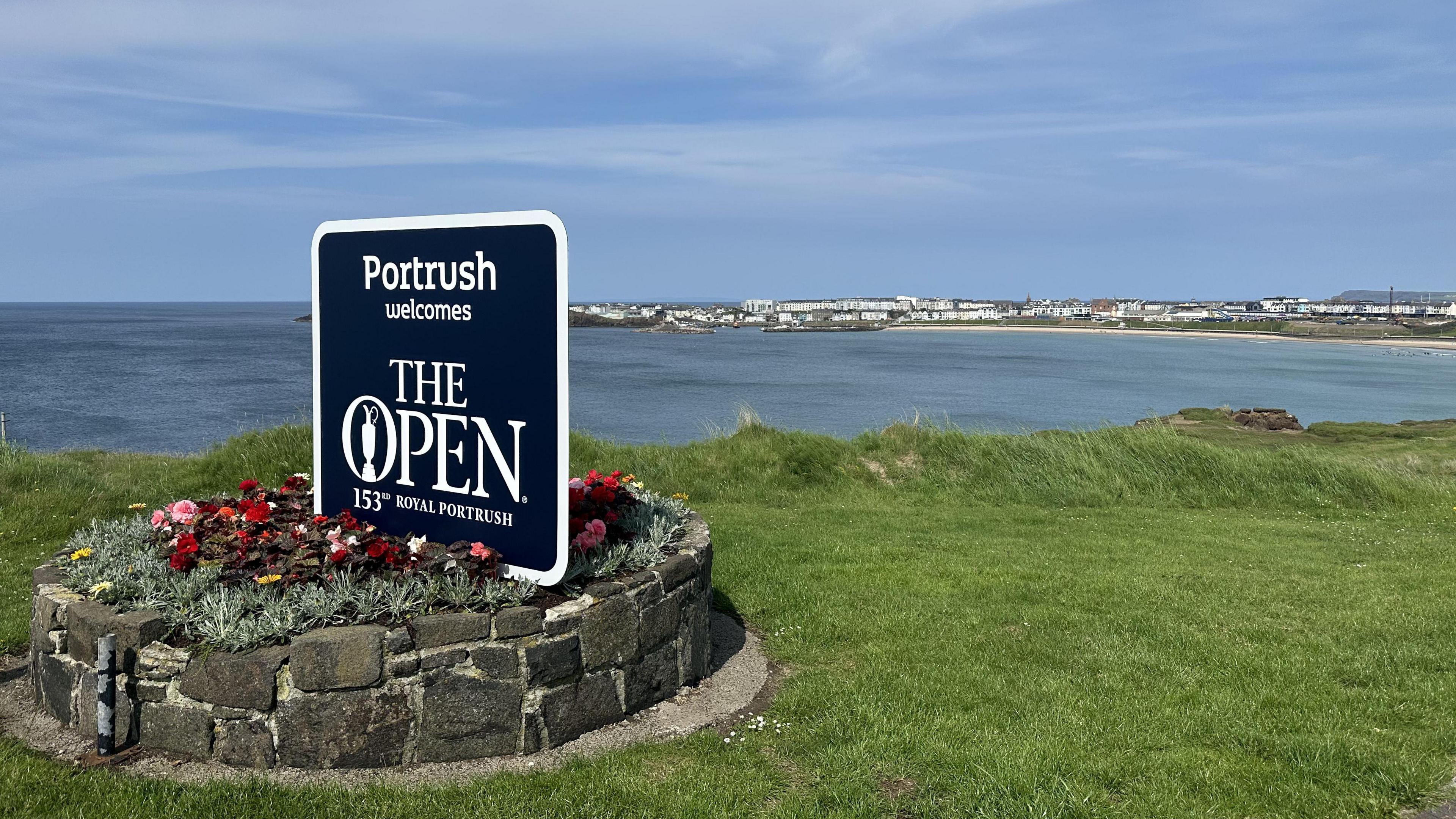 A sign which reads "Portrush welcomes The Open" is placed on a grassy hilltop overlooking the water with Portrush in the background. There are blue skies and the sign is placed in a rockery with flowers.