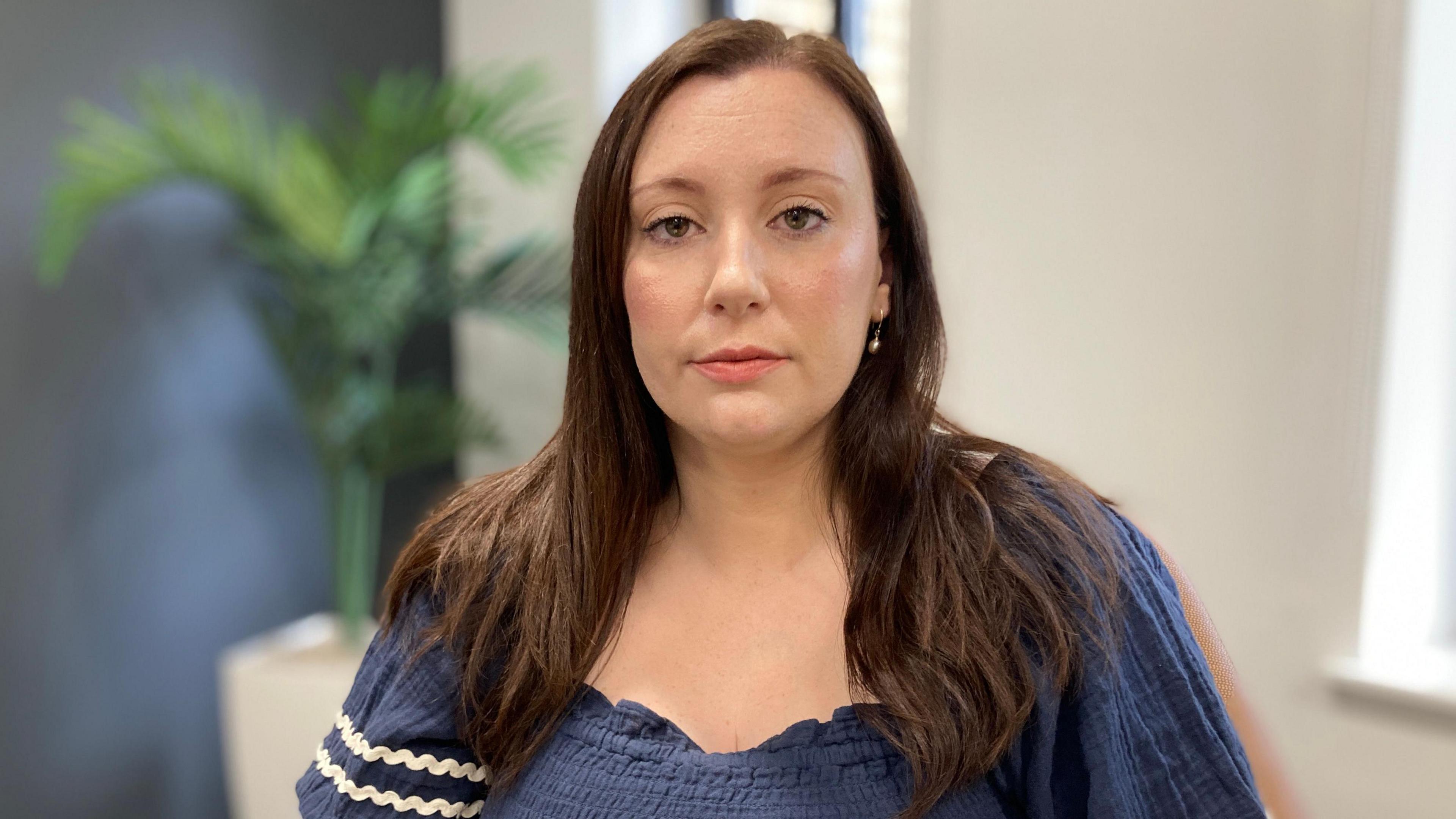 A woman with long brown hair is looking at the camera. She is wearing a blue top and behind her is a plant.