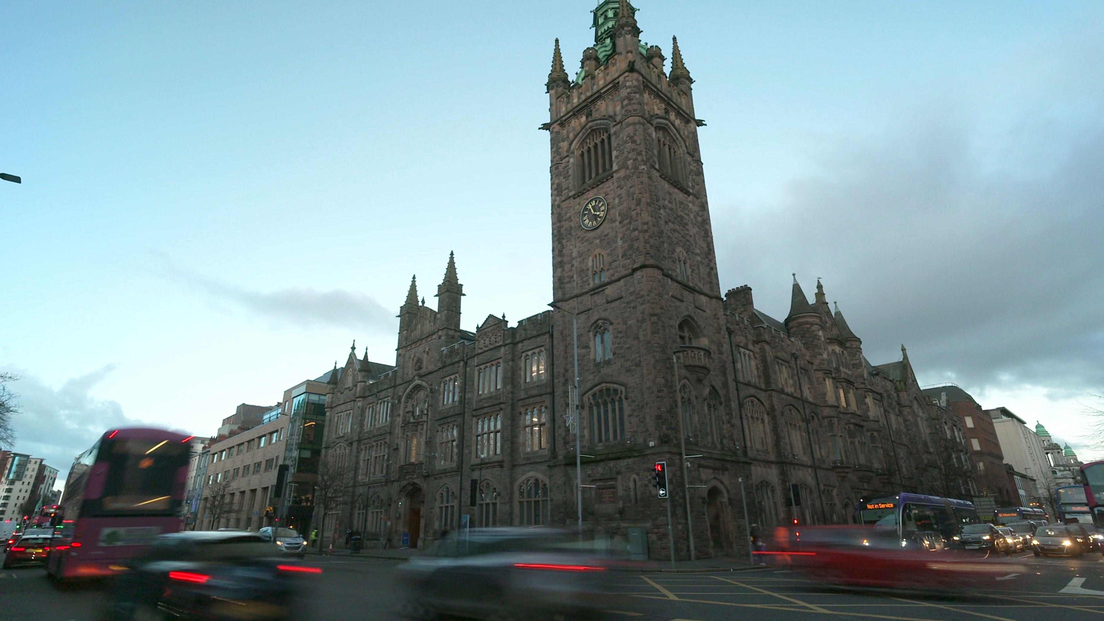 A large church building in Belfast city centre shows, stained glass windows and a bell tower, bearing a gold and black clock. Behind the building is a gloomy, cloudy sky. Traffic is blurred as it passes the camera.