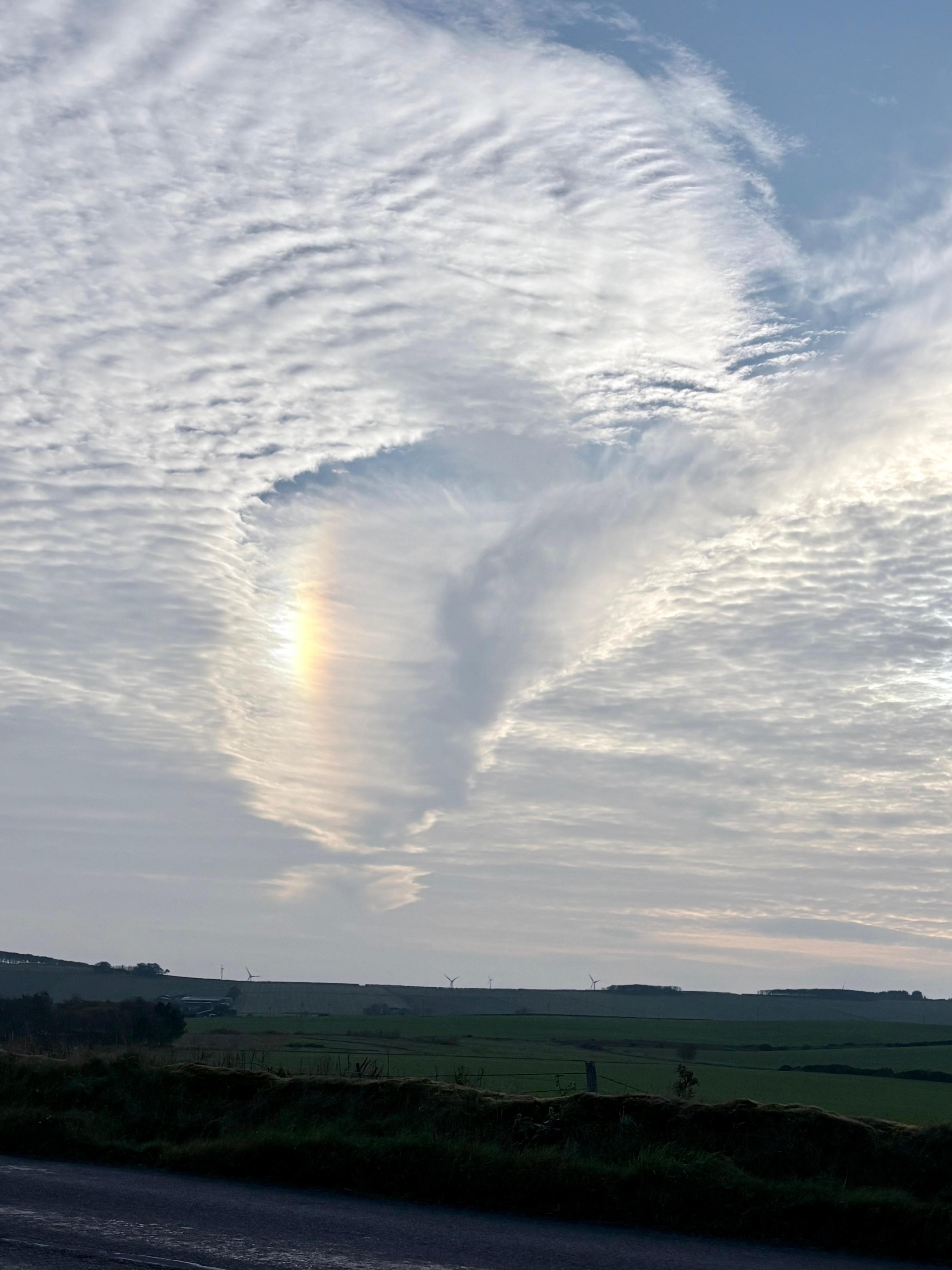 A dramatic fallstreak hole cuts through a blanket of white cloud above open farmland near Turriff, Aberdeenshire. A faint rainbow-coloured patch of light appears along the edge of the gap, while the landscape below shows green fields, hedgerows and distant wind turbines beneath the bright sky.