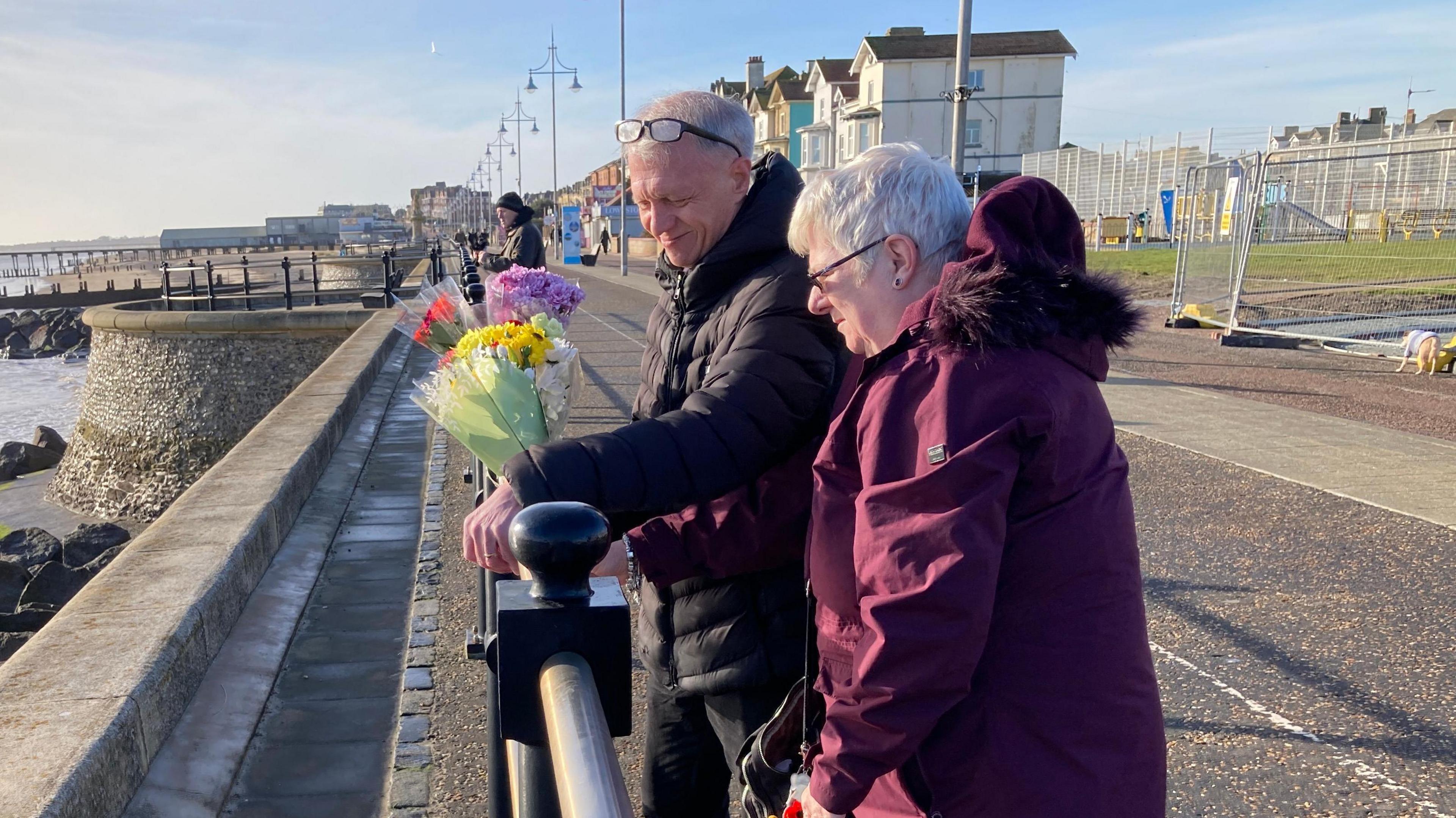 Tricia and Barry Butler are pictured placing flowers on a black railing on Lowestoft seafront. They both have short grey hair and glasses. Mrs Butler wears a maroon coloured coat, while Mr Butler wears a black coat.