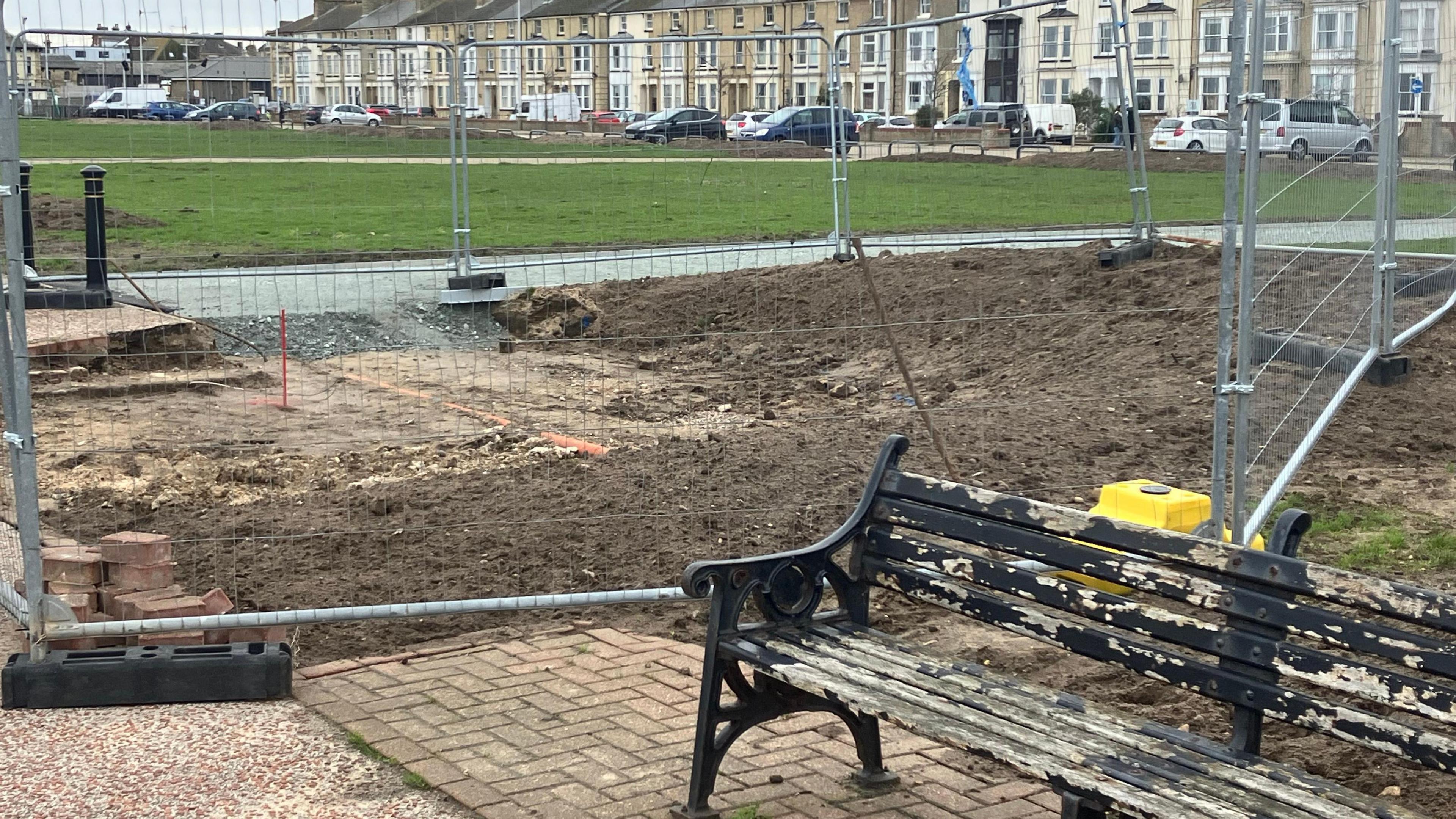 In the foreground, a weathered, black bench sits on brick paving. There is metal fencing behind it and beyond the fencing, the ground has been dug up. In the distance, the other side of a green, there are terraced houses. 