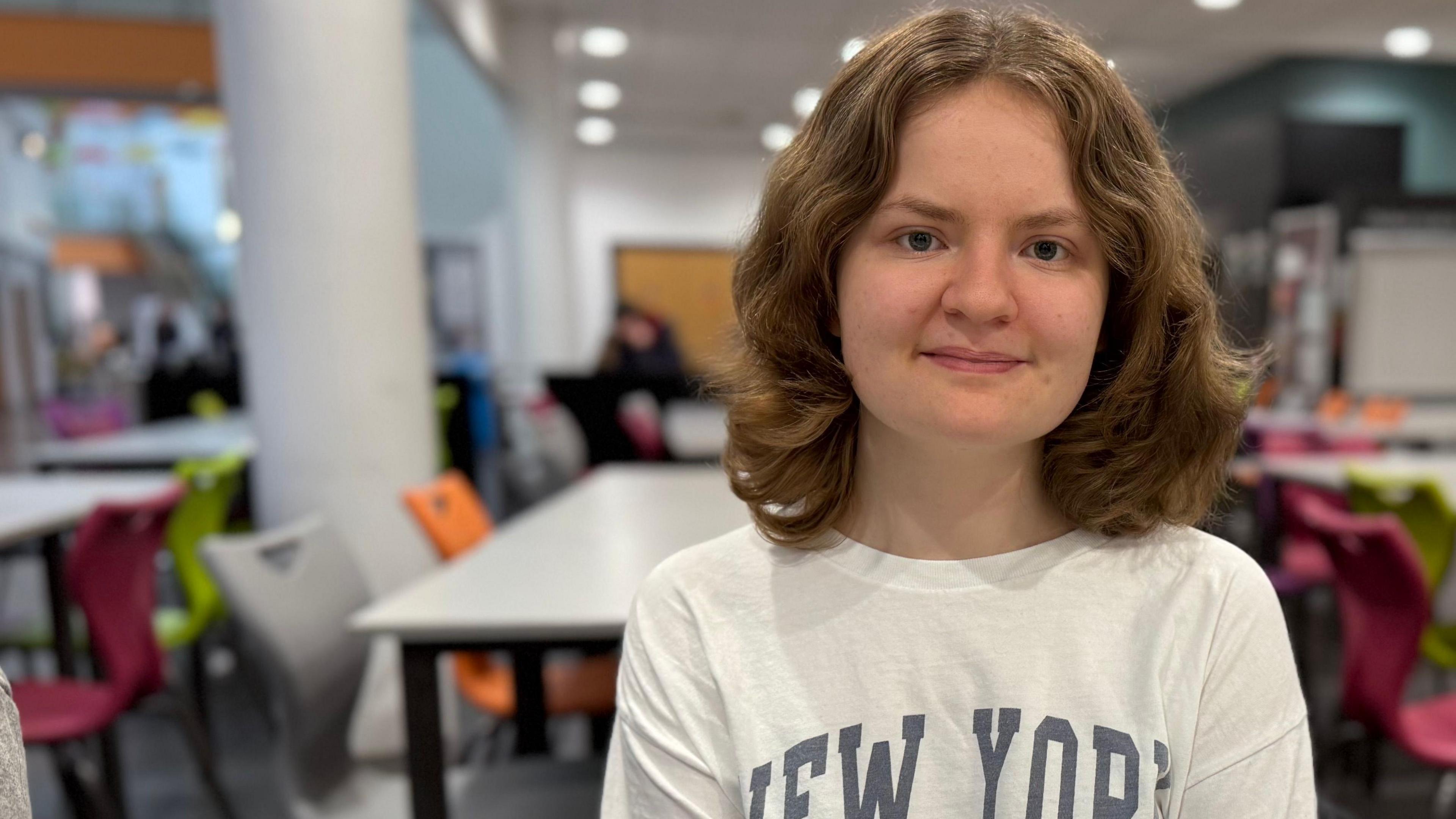 Elinor Lewis sits at a table within a food canteen at Coleg y Cymoedd in Nantgarw, she wears a long sleeve white t-shirt with the words New York on it. She smiles at the camera. She has mousy brown shoulder-length hair and brown eyes. 